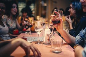 People at a restaurant table, drinking wine and talking. Pink tablecloth, soft lighting, and glasses.