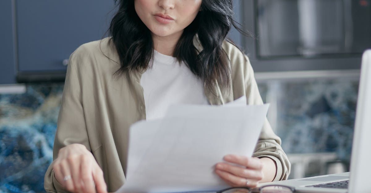 A woman is sitting at a table holding a piece of paper in front of a laptop computer.
