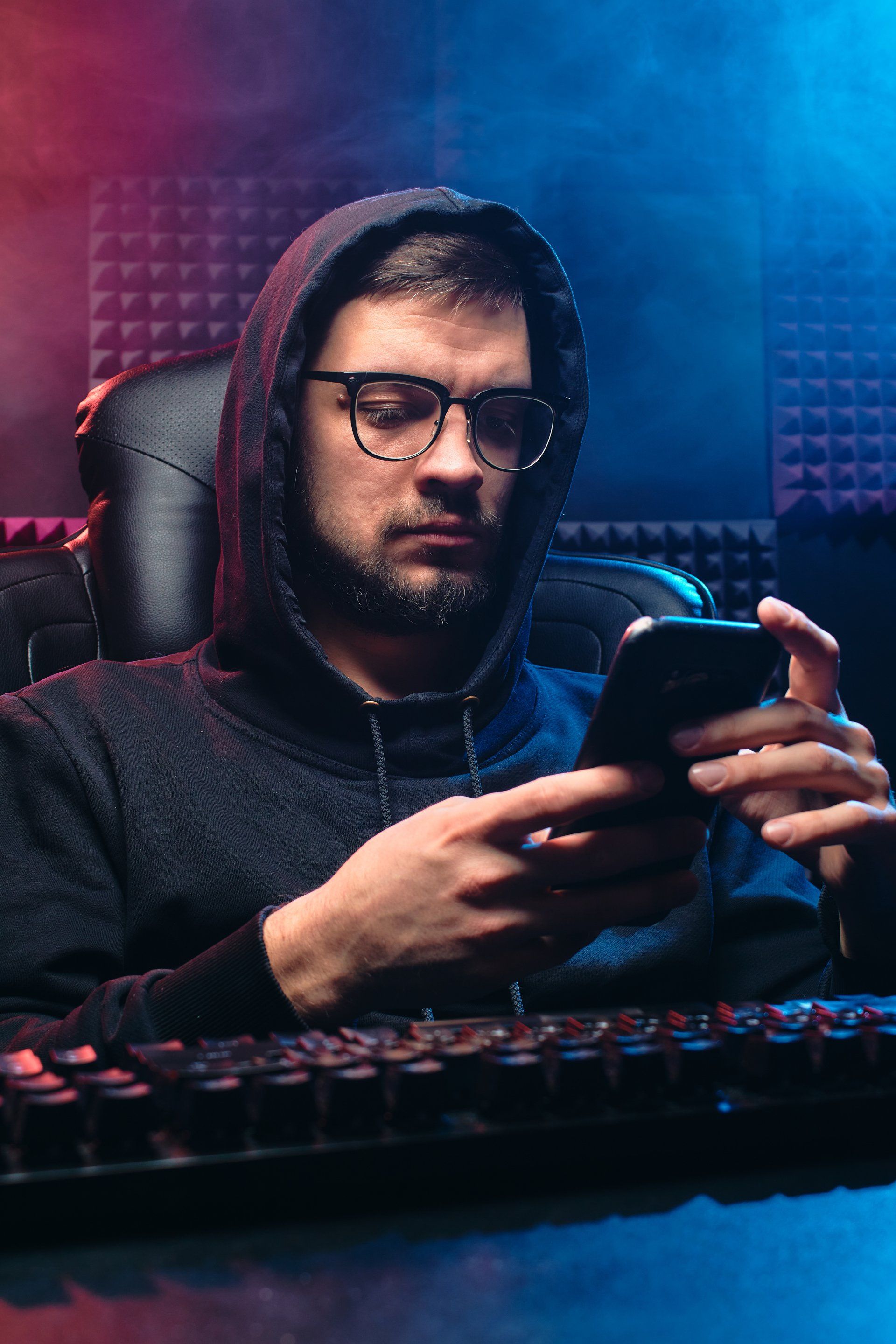 A man in a hoodie is sitting at a desk using a cell phone.