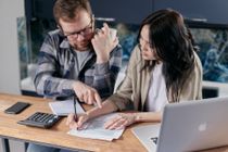 A man and a woman are sitting at a table looking at papers and a laptop.