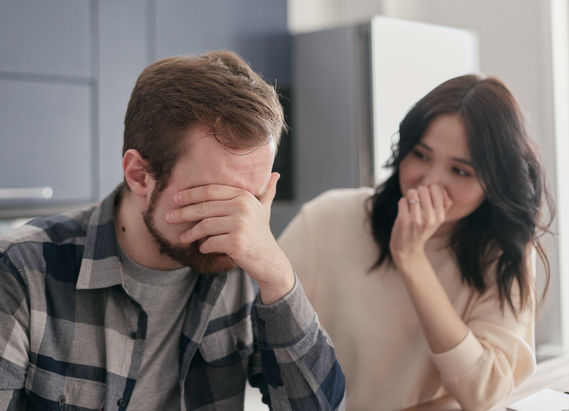 A man is covering his face with his hands while a woman laughs.