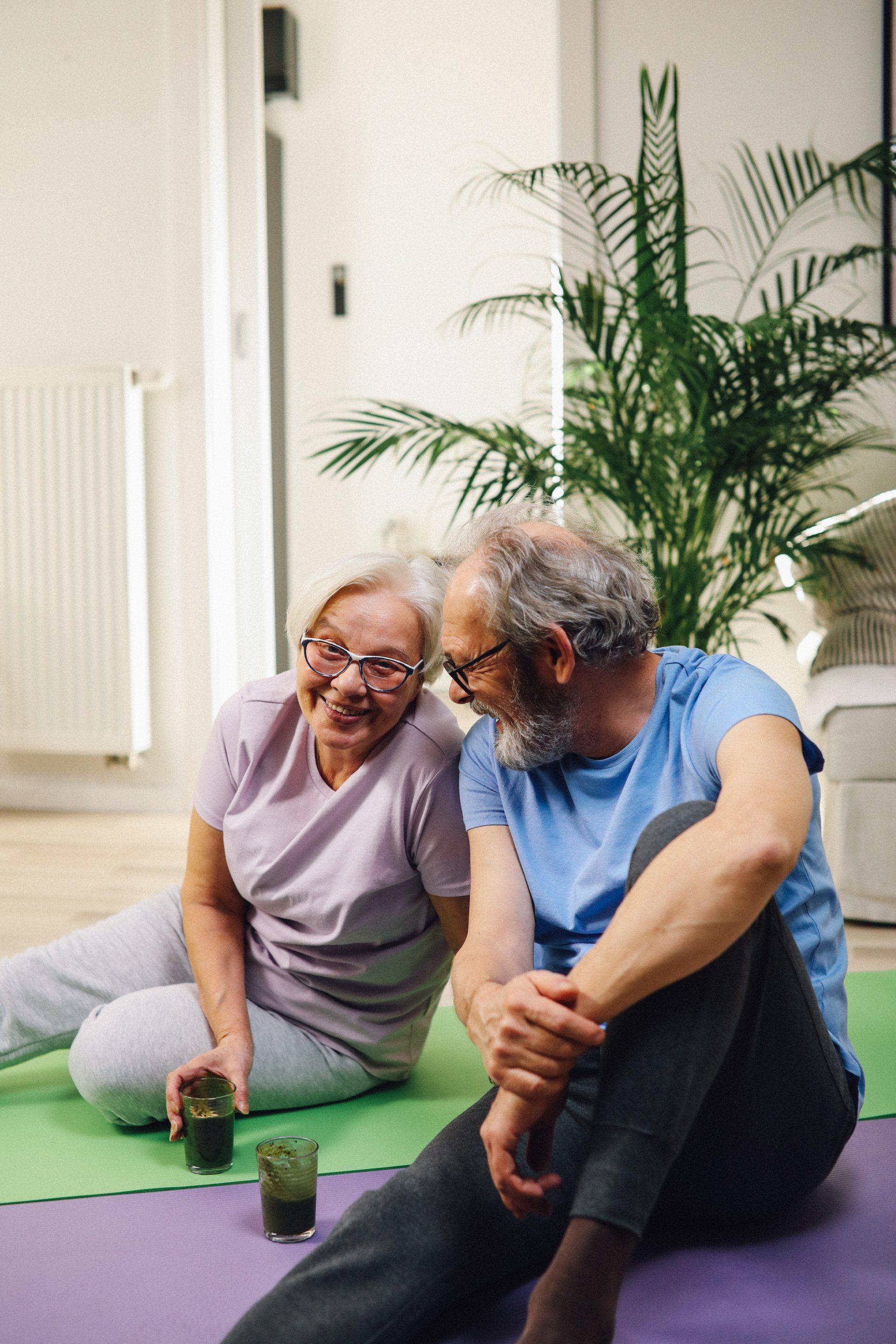 An elderly couple is sitting on a yoga mat in a living room.