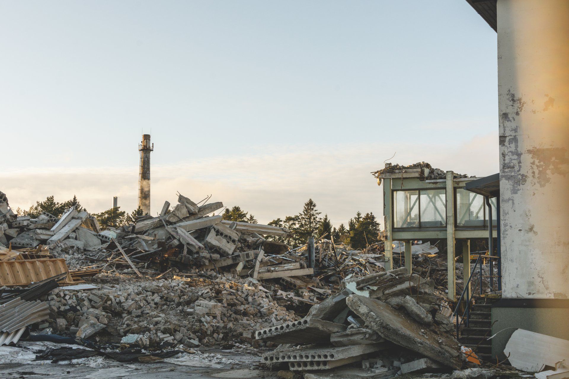 Demolished building site with rubble, partially standing structure, and a distant smokestack. Overcast sky.