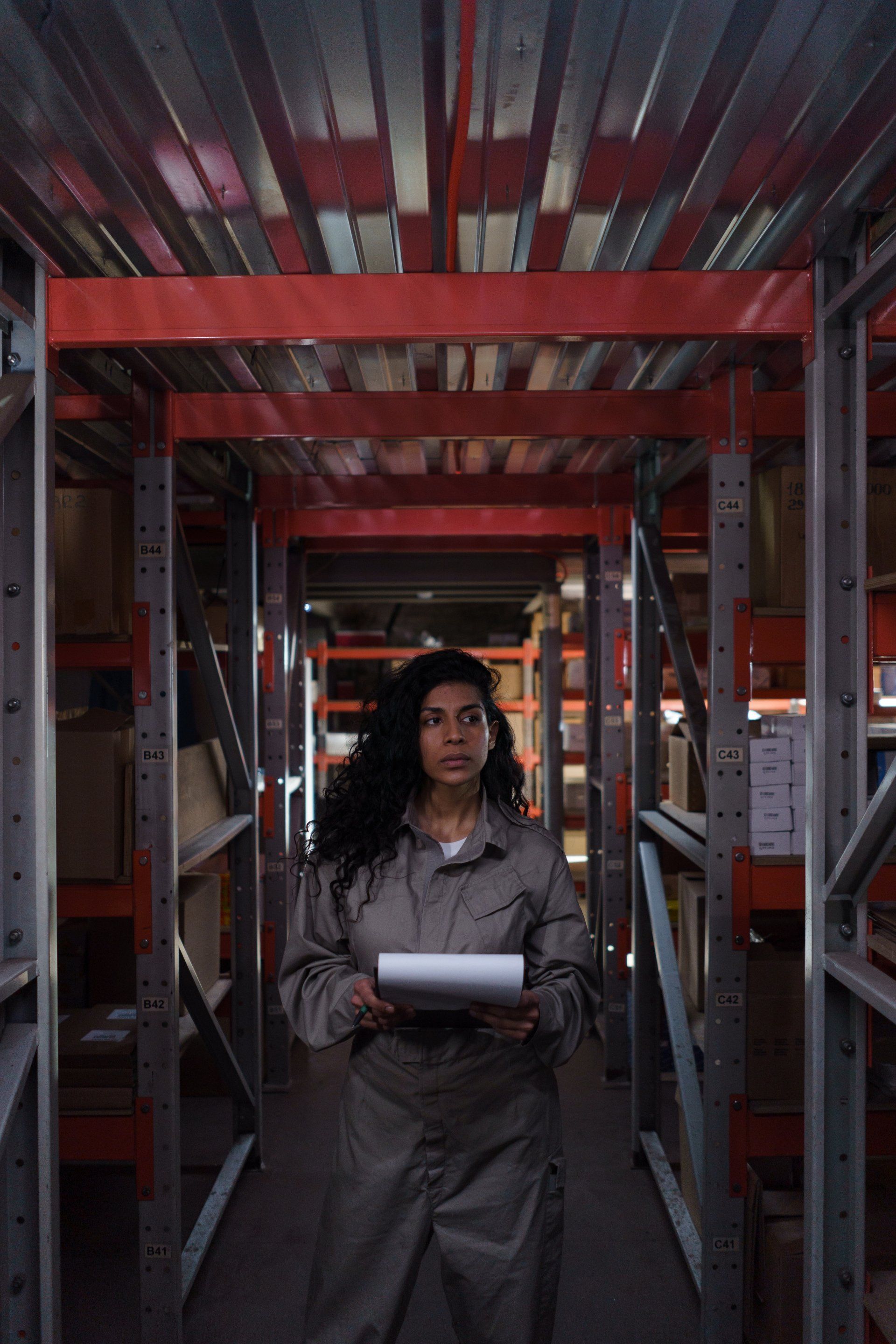 Woman in jumpsuit in a warehouse, holding a clipboard, looking around.