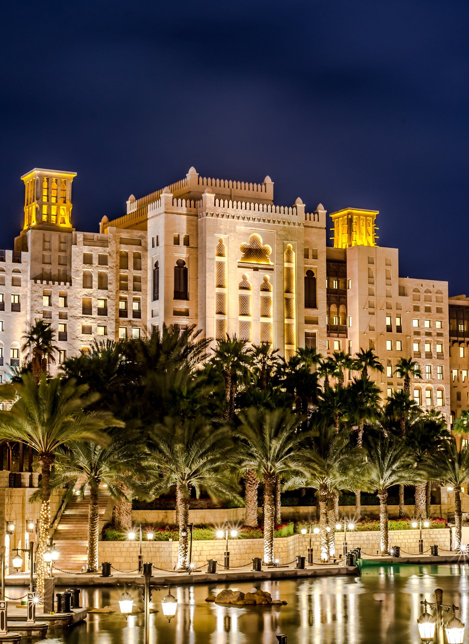A large hotel is lit up at night with palm trees in front of it.