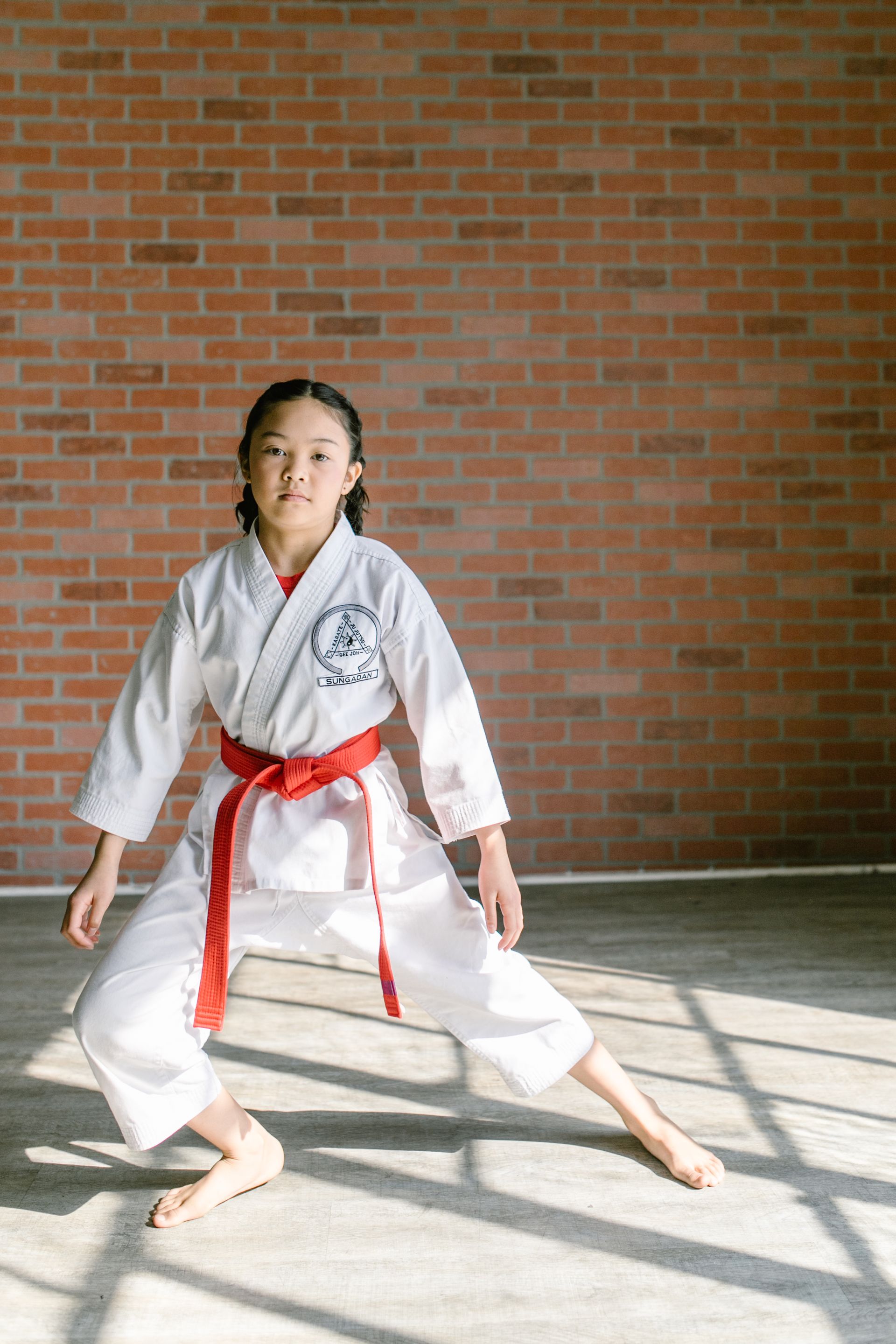 Een jong meisje in een wit karate-uniform met een rode riem staat voor een bakstenen muur.