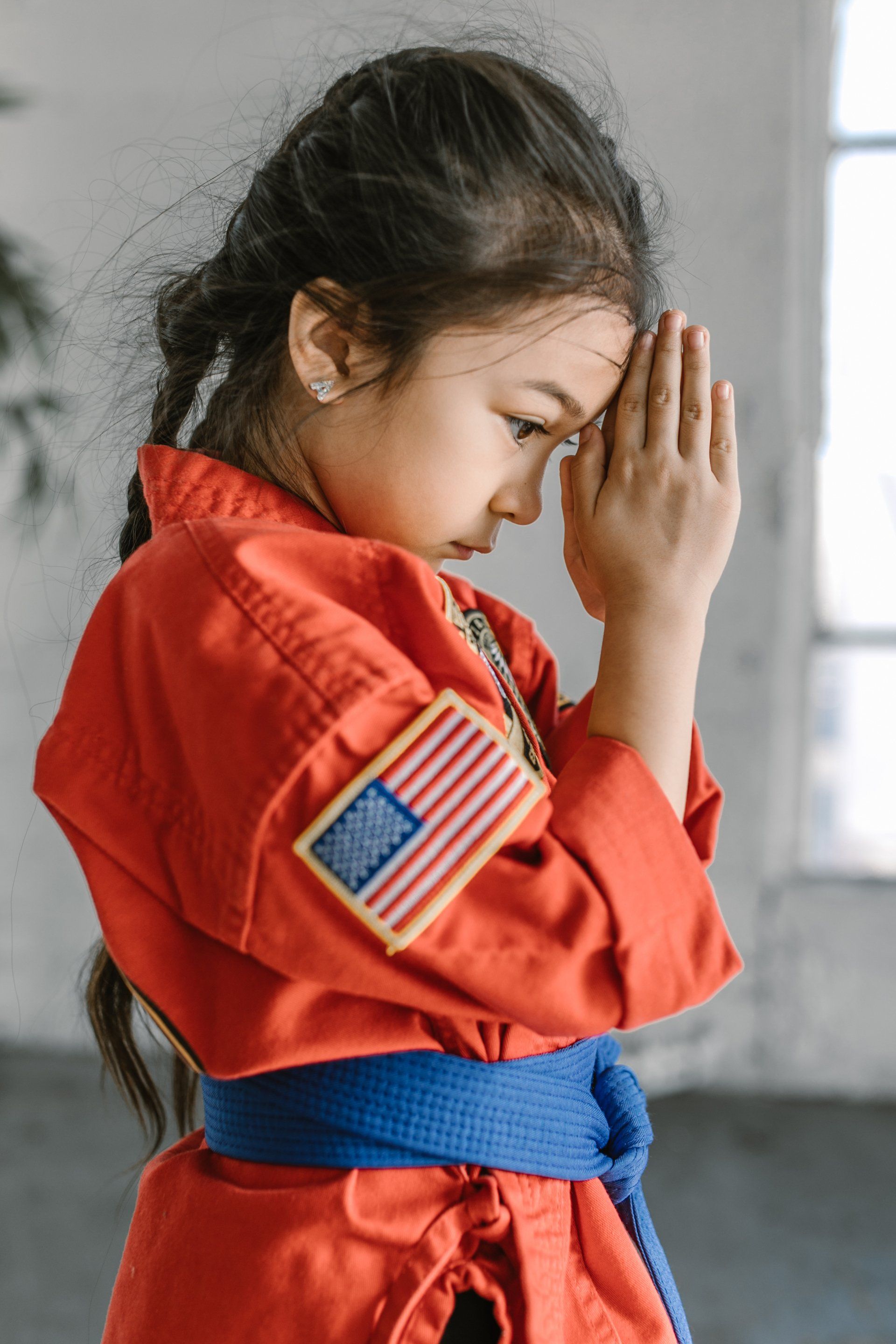 A young girl wearing a red karate uniform and a blue belt is praying.