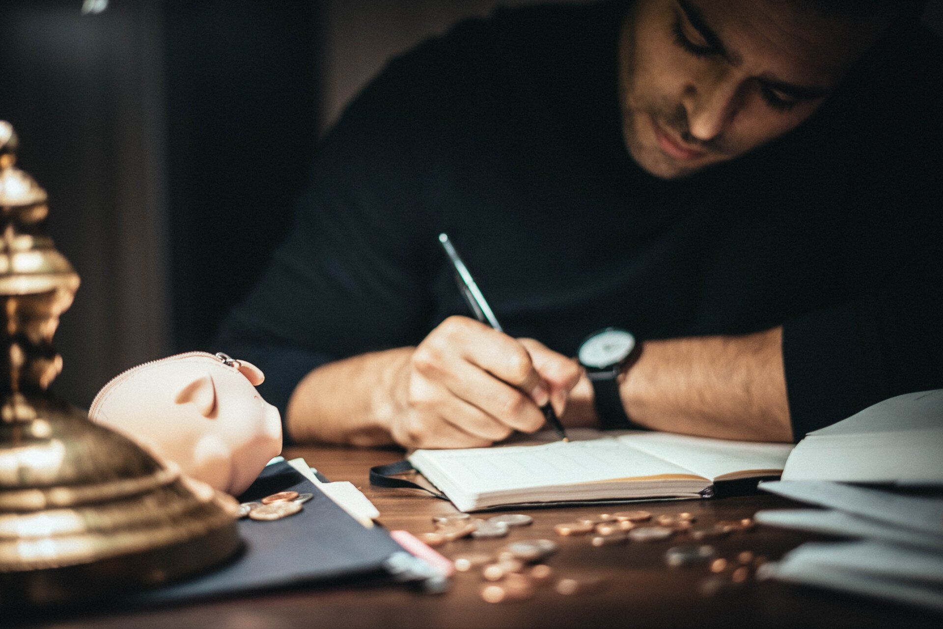 a man is sitting at a table writing in a notebook next to a piggy bank .