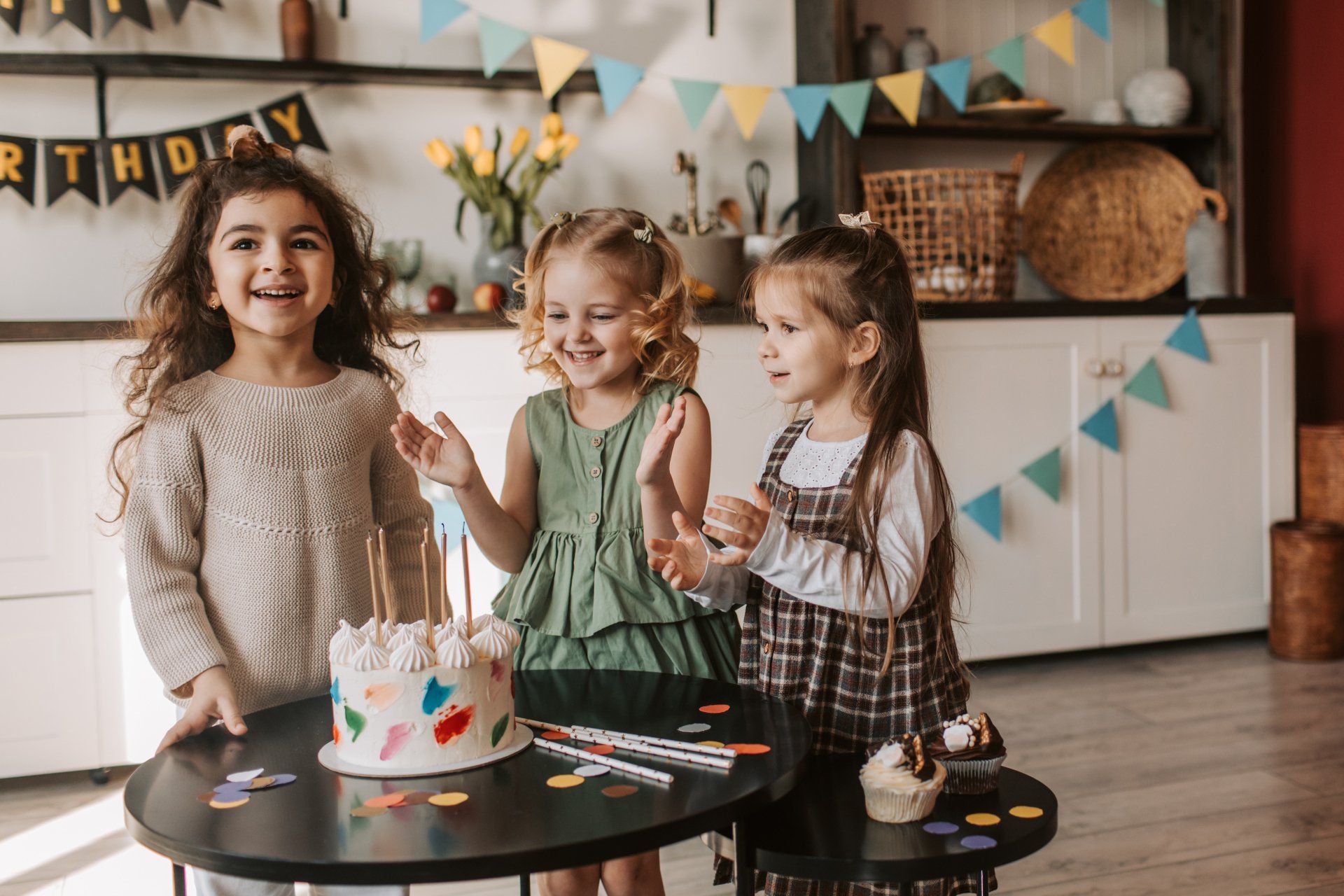 Three young Jewish girls smiling & standing at a table with a birthday cake on it