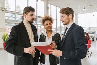 A group of business people are standing in a car showroom looking at a clipboard.