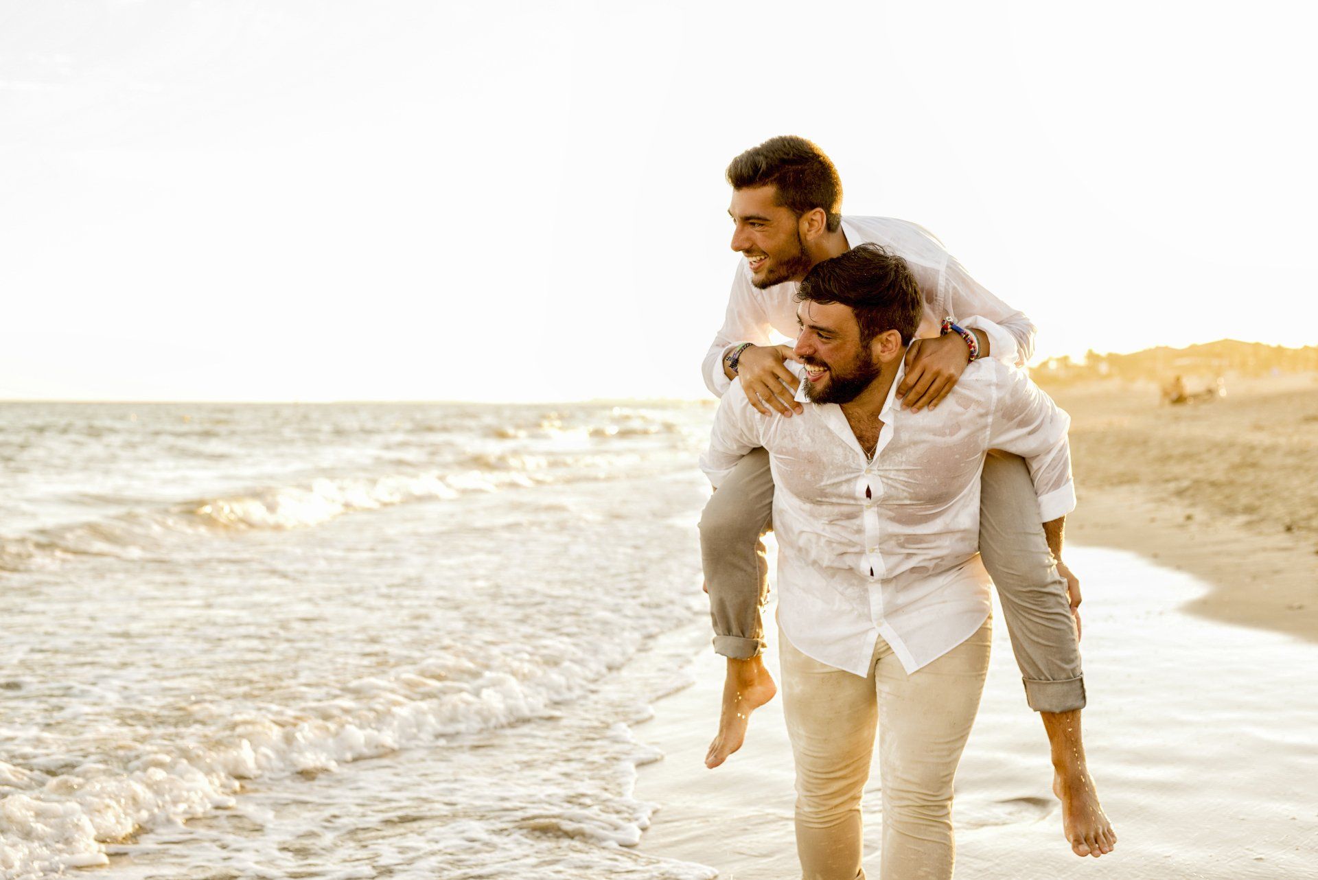 Two men in wet, white shirts on a beach; one giving the other a piggyback ride. They smile, backlit by the sun, near the water's edge.