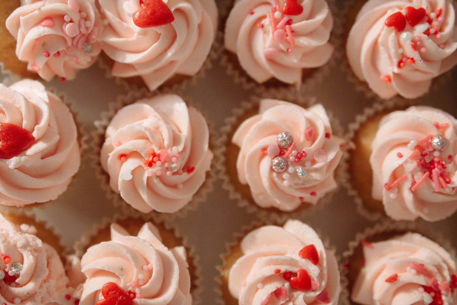 Pink frosted cupcakes decorated with sprinkles and heart-shaped candies in a bakery box.