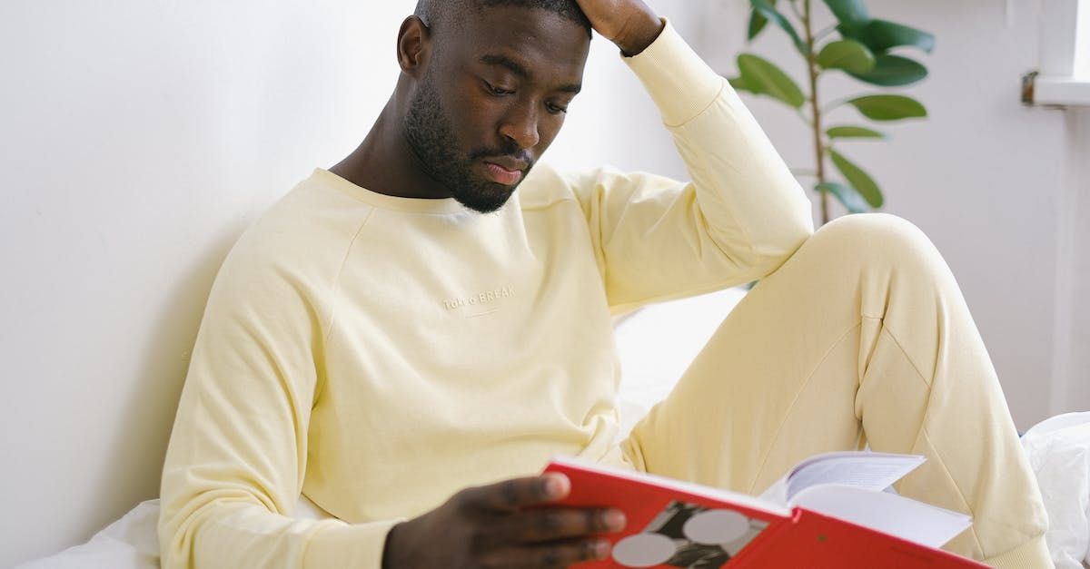 A man is sitting on a bed reading a book.
