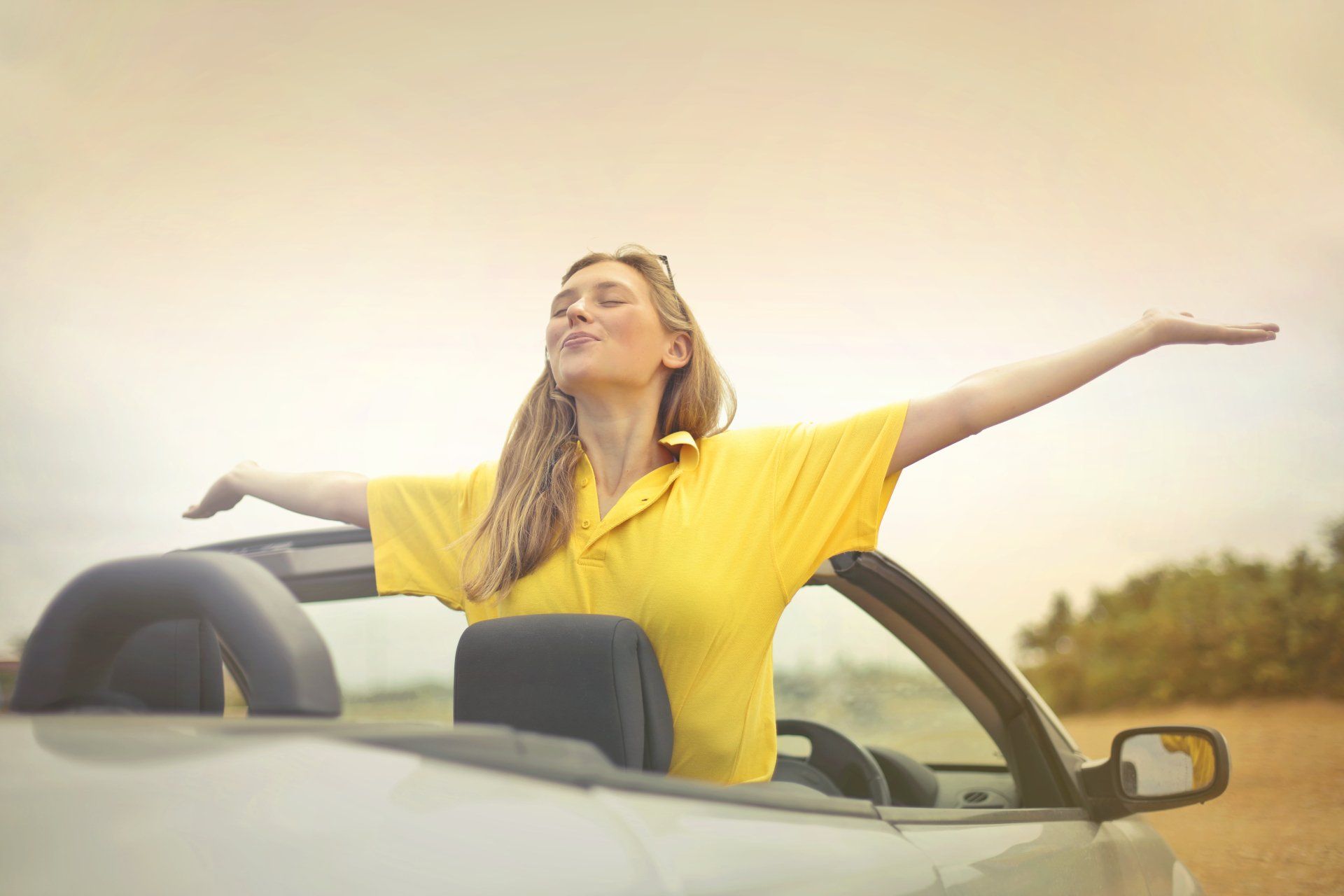 A woman in a yellow shirt is sitting in a convertible car with her arms outstretched.