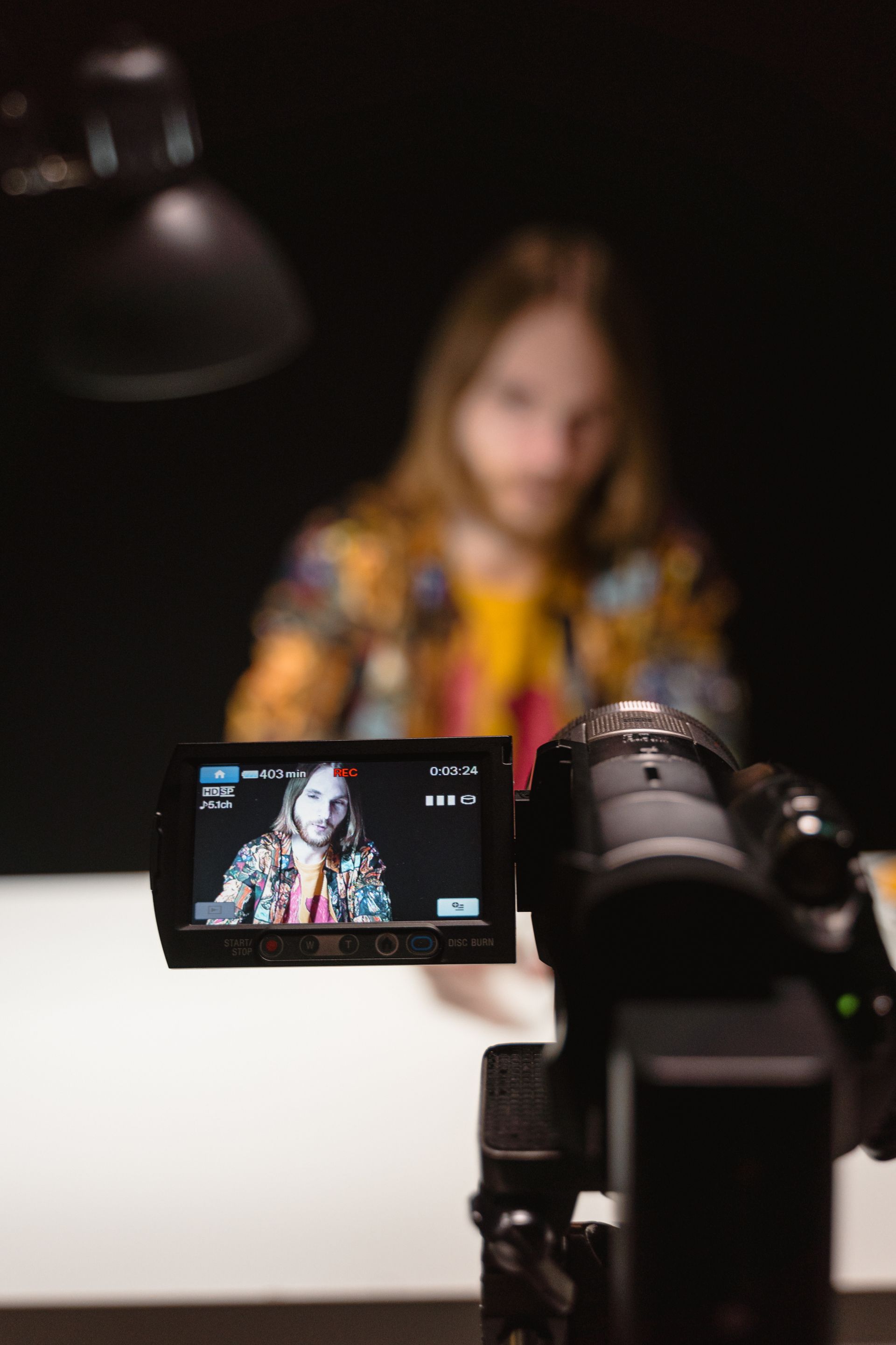 Person with long hair filming a video. Camera display shows him, vibrant clothing, dark background.