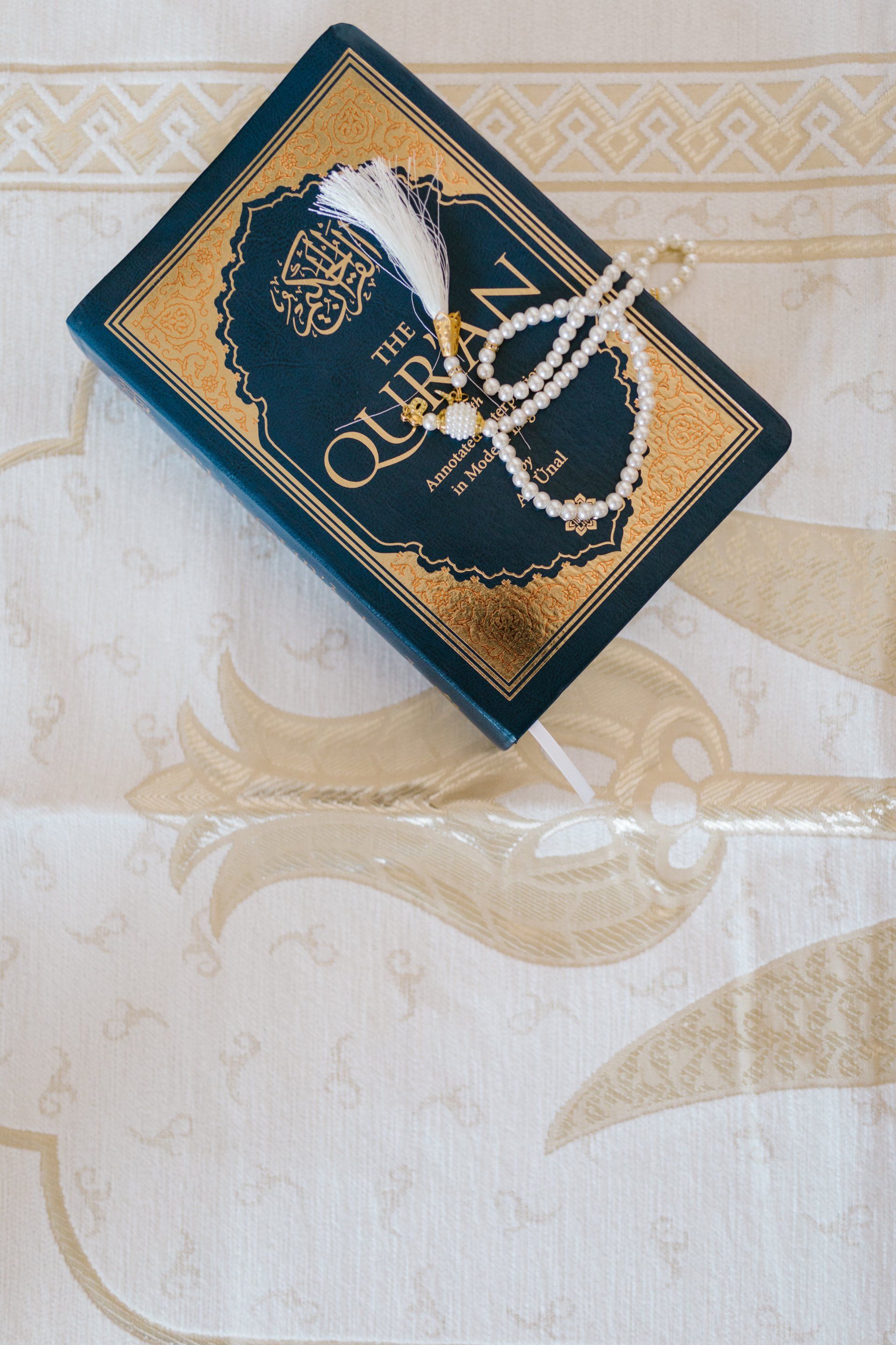 Blue Quran book with gold trim and white prayer beads on a patterned cream surface.