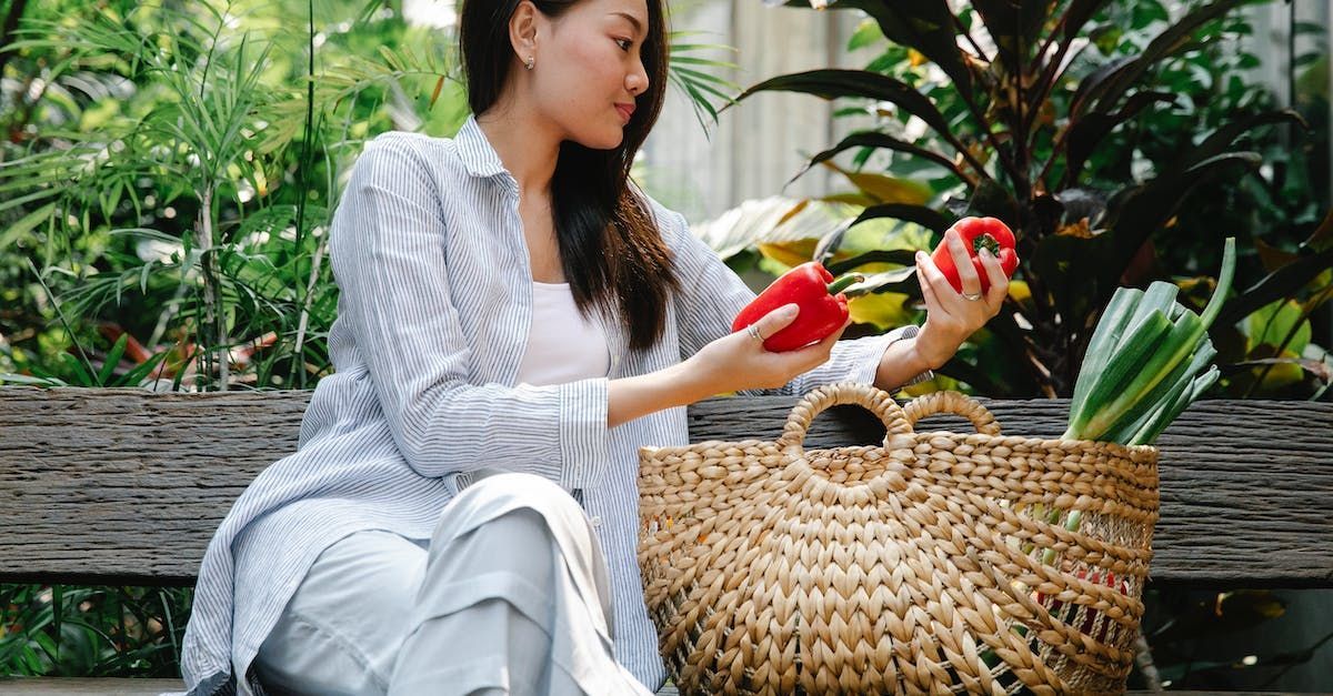 a woman is sitting on a bench holding a red pepper next to a basket of vegetables .