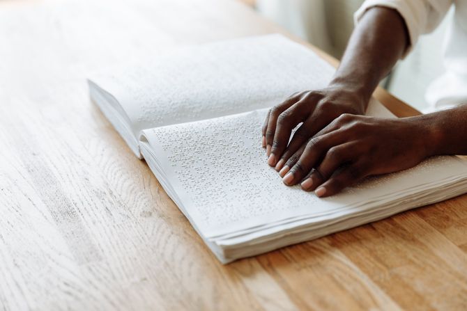 Des mains posées sur un livre ouvert avec du texte en braille, sur une table en bois.