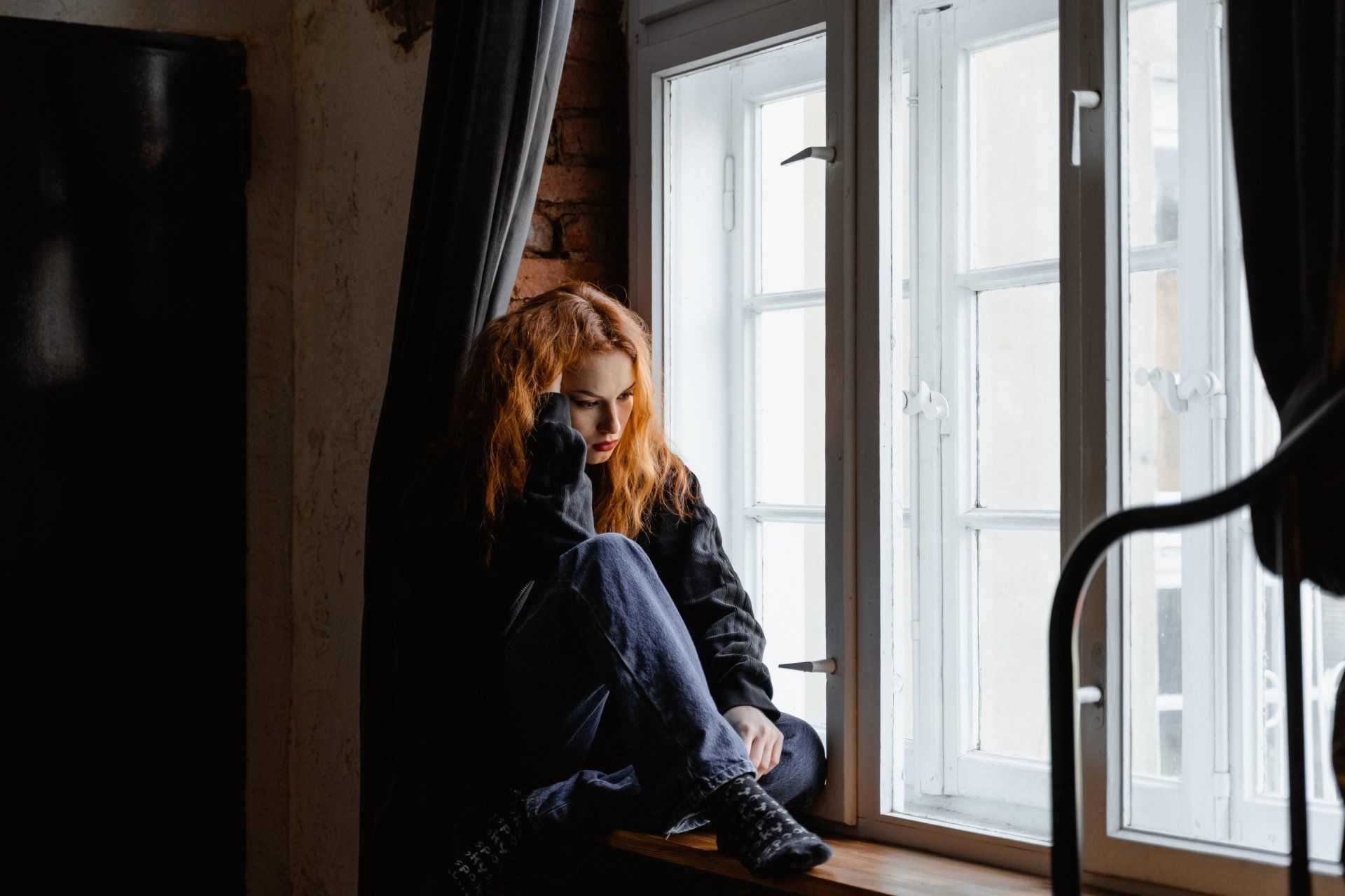 A distraught woman sitting by a window, her gaze heavy with the emotional burden of coping with a family member's hoarding behavior.