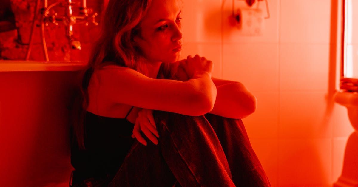 Woman in red light, sitting on the floor, knees to chest, looking sad.