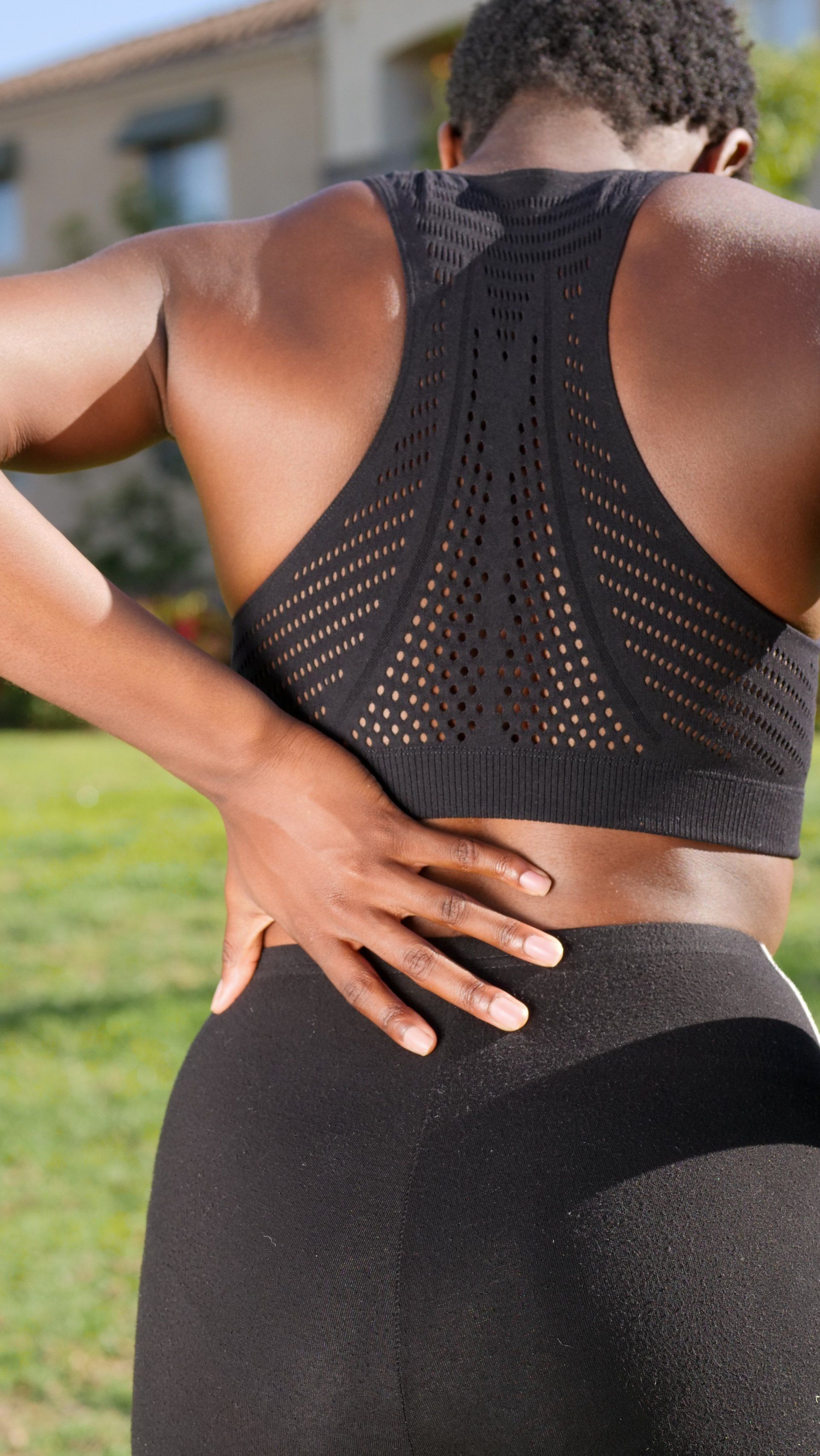 A woman in a black sports bra and leggings is holding her back in pain.