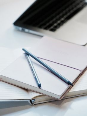 A notebook with two pens on top of it next to a laptop