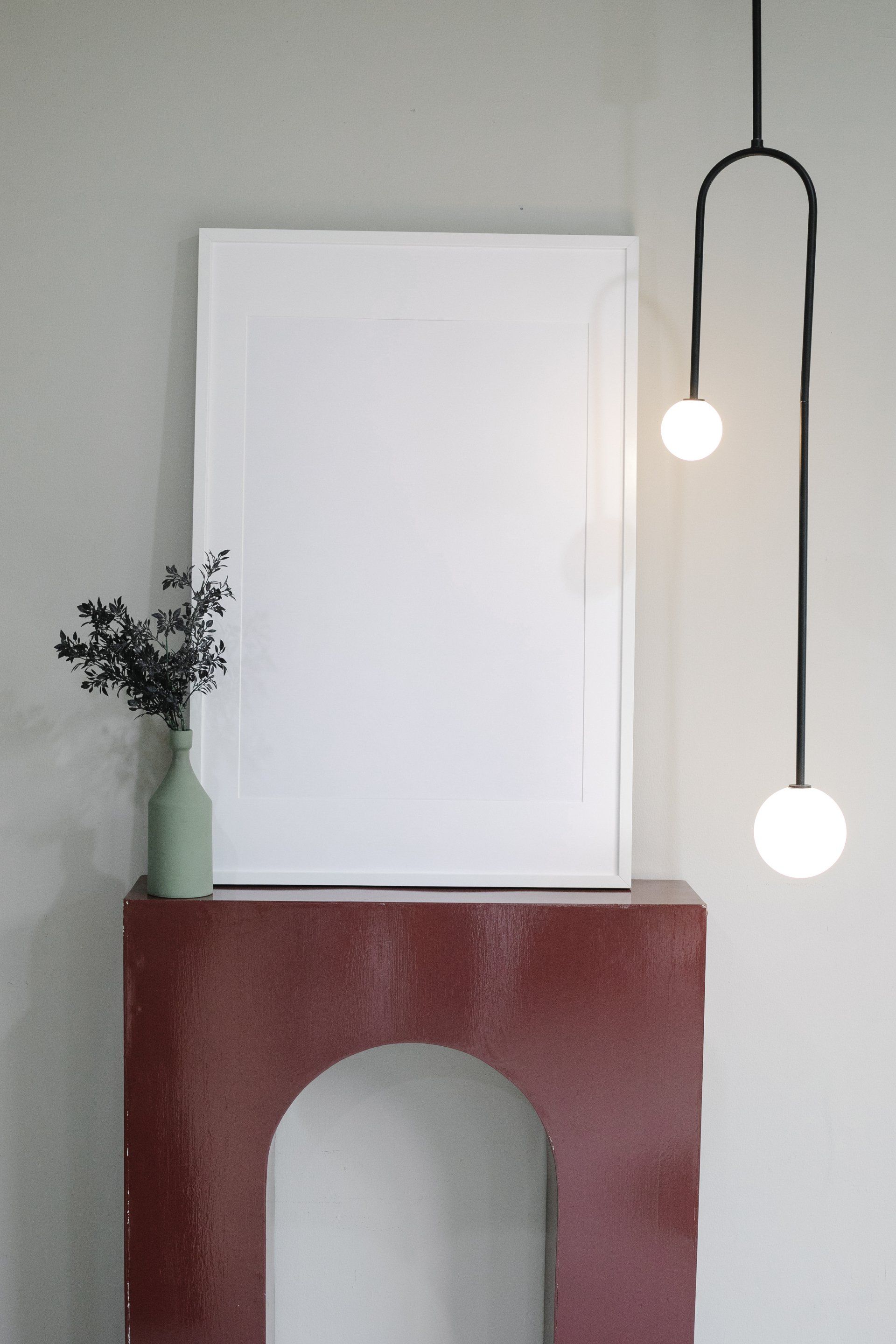 Red arch-shaped shelf holding blank canvas and vase with plant, lit by a two-bulb pendant lamp.