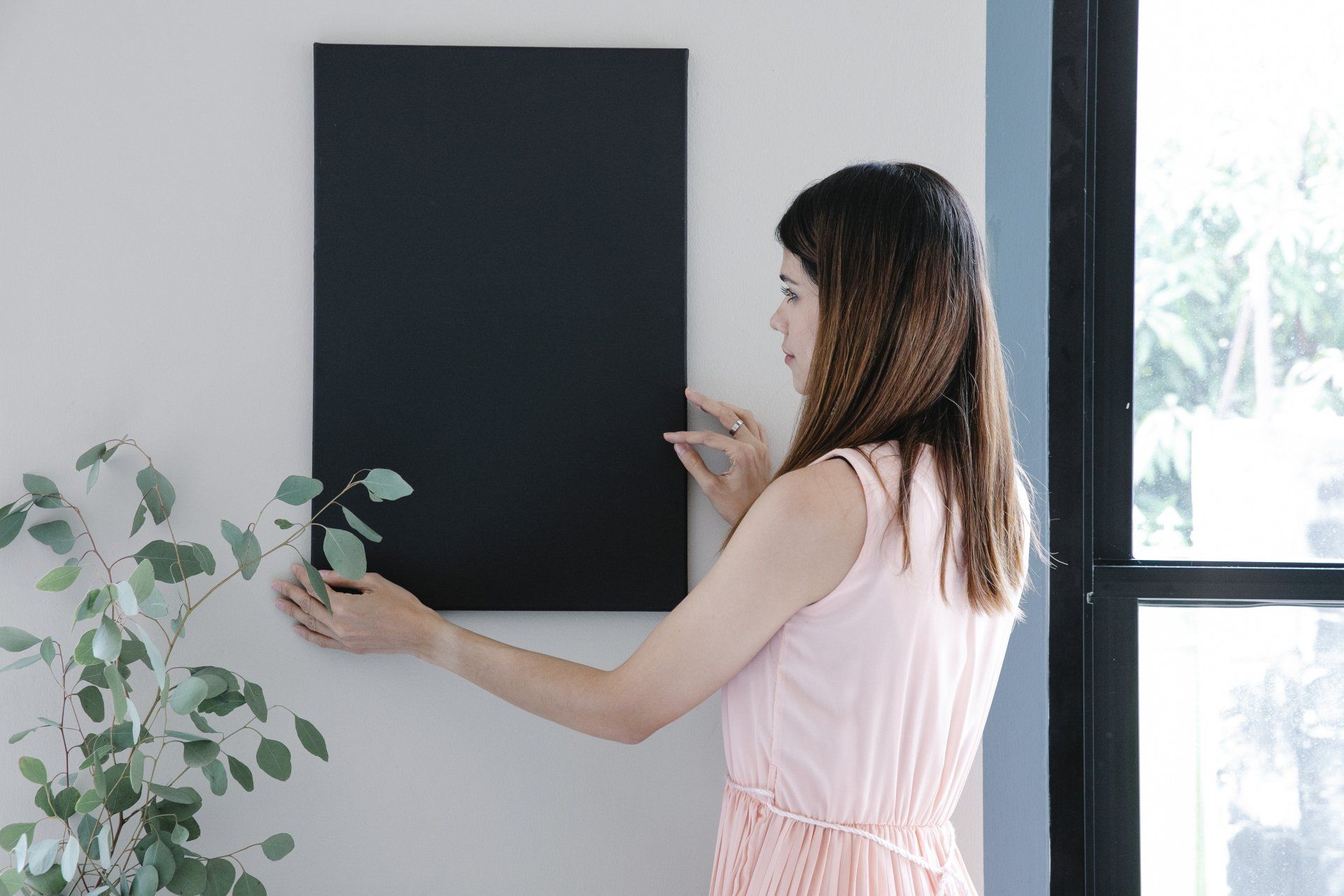 A woman in a pink dress is hanging a blackboard on a wall.