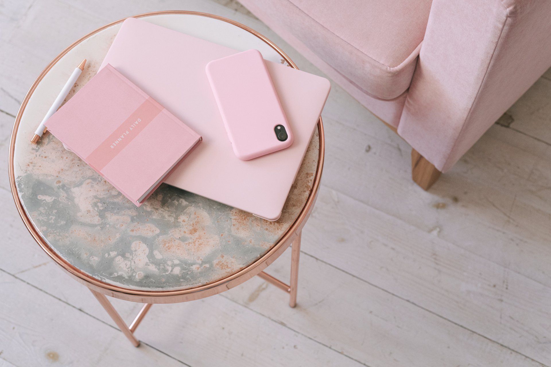 Pink laptop, phone, and notebook on a small table next to a pink chair on a wooden floor.