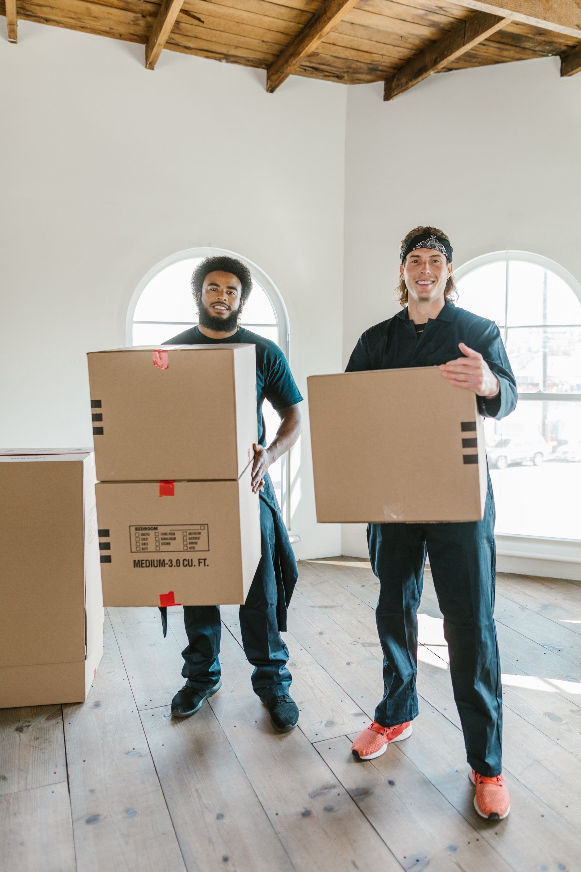 Two men are carrying cardboard boxes in an empty room.