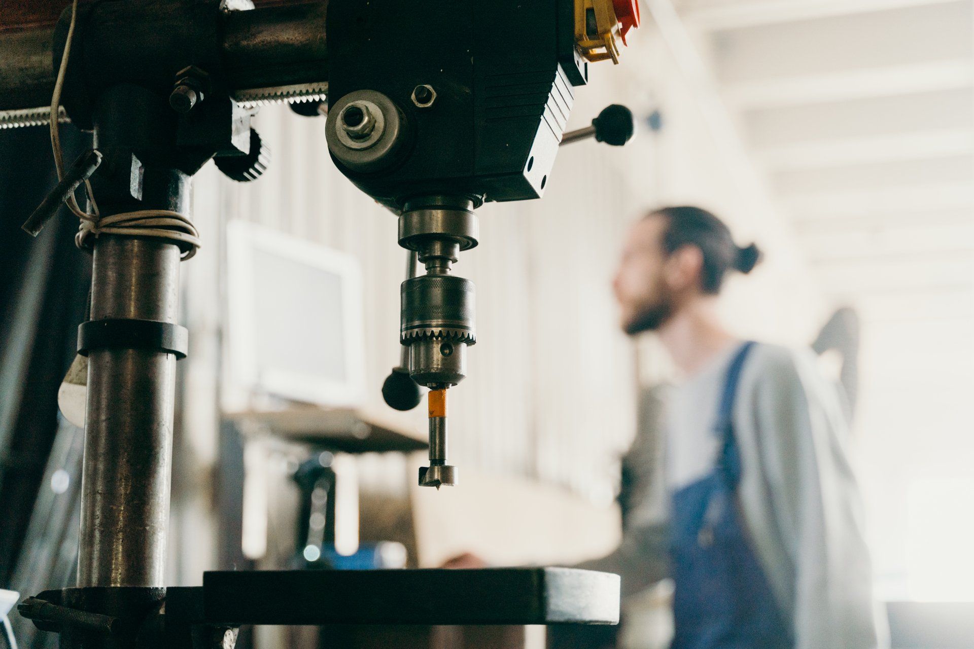 A man in overalls looks at a drill press in a workshop.