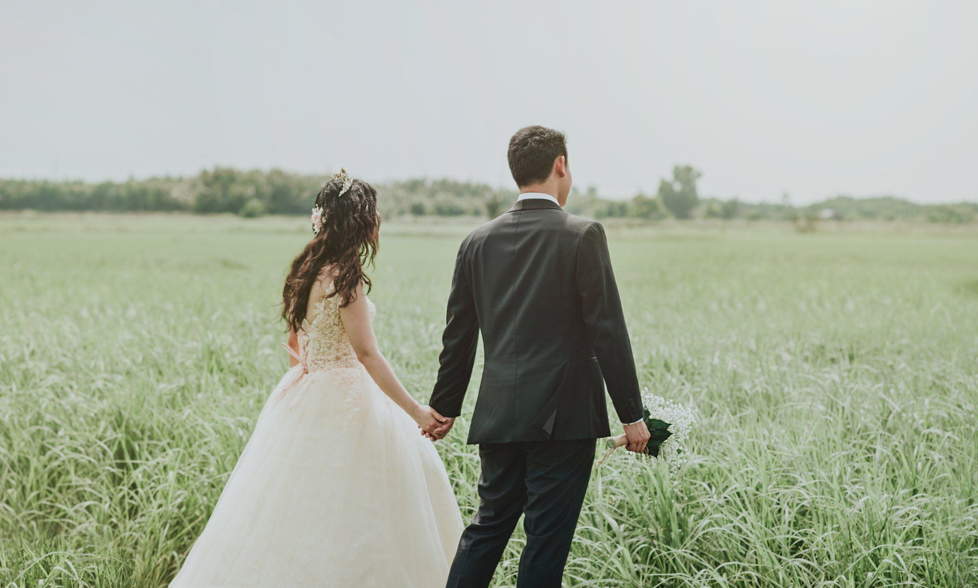 A bride and groom are walking through a grassy field holding hands.