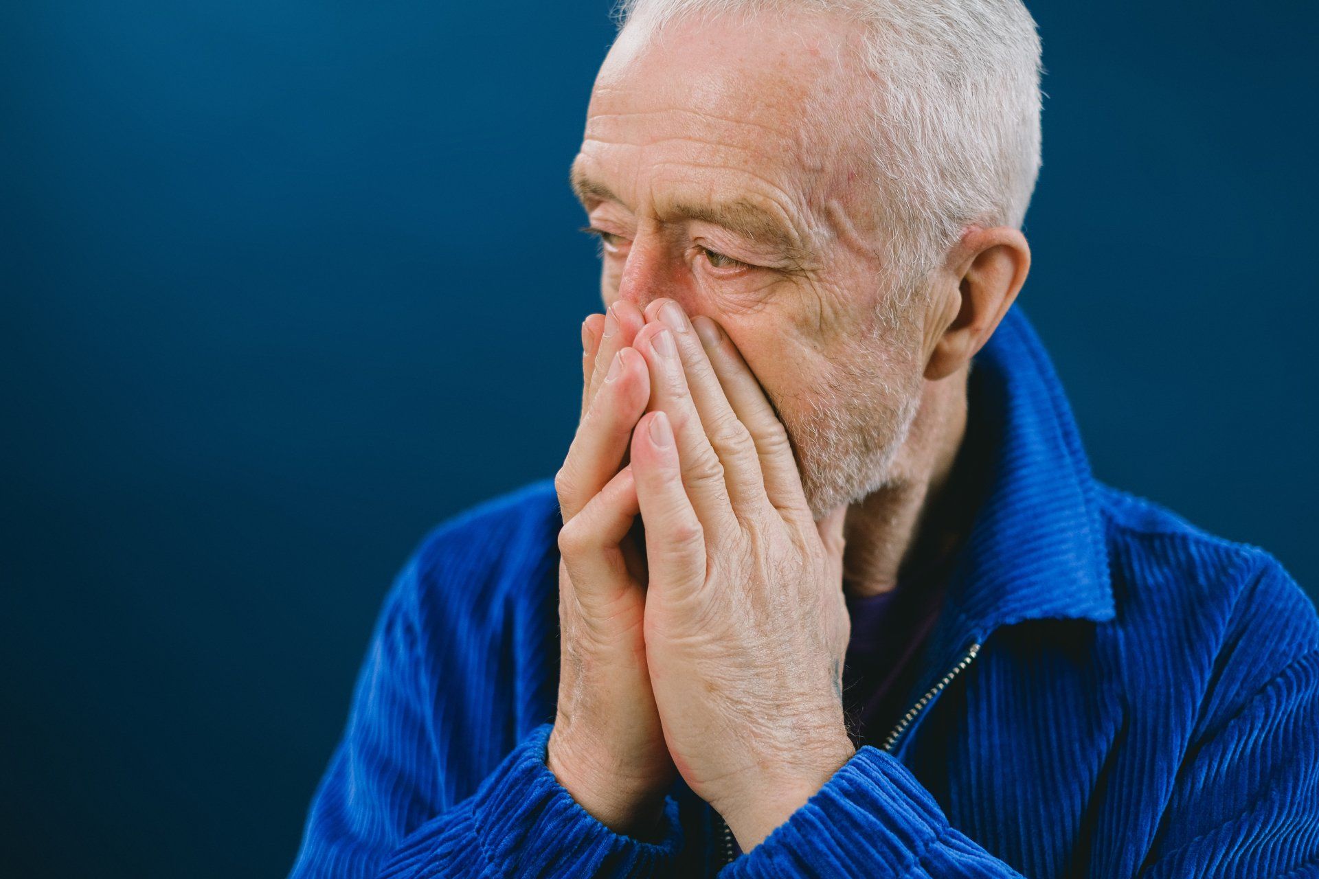 An elderly man in a blue jacket is covering his mouth with his hands.