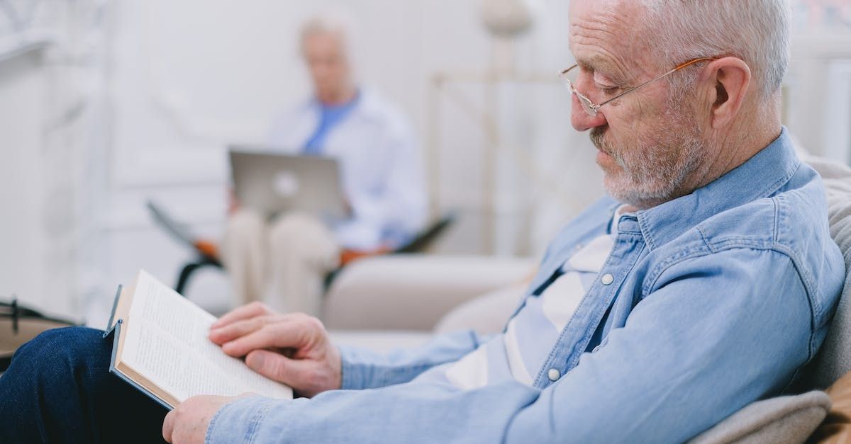 an elderly man is sitting on a couch reading a book .