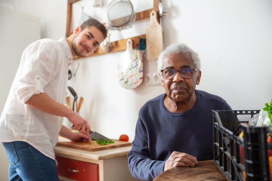 A young man is cutting vegetables for an older man in a kitchen.