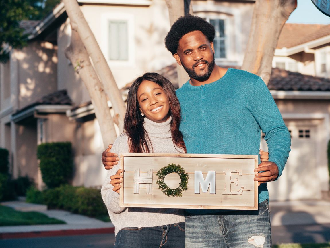 a man and a woman are standing in front of a house holding a sign that says `` home '' .