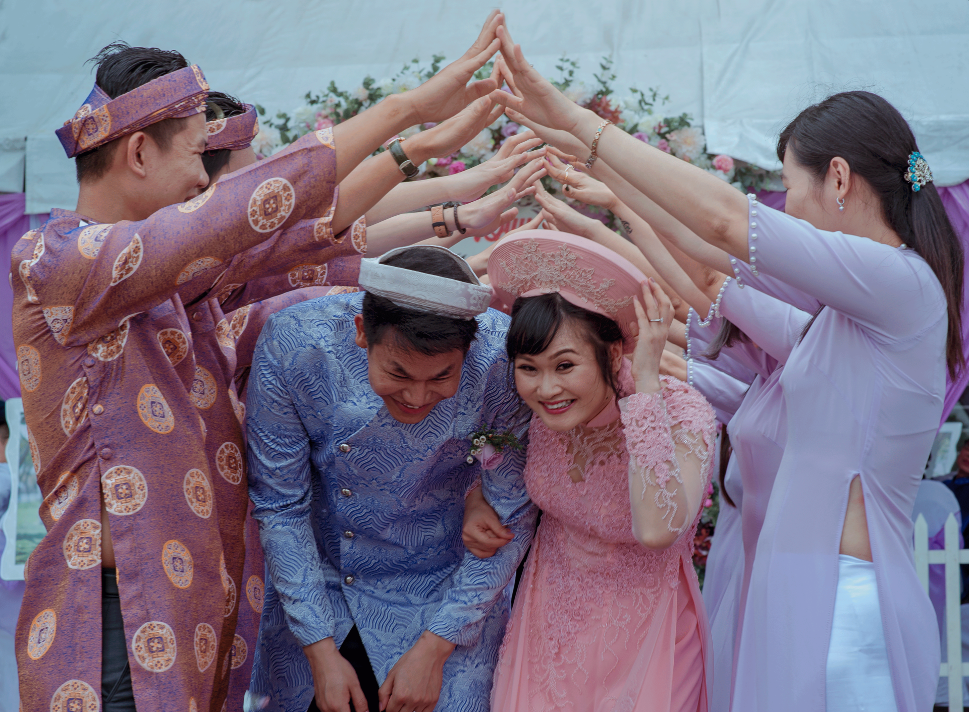 An Asian couple with hats on going under the outstretched arms of their wedding guests