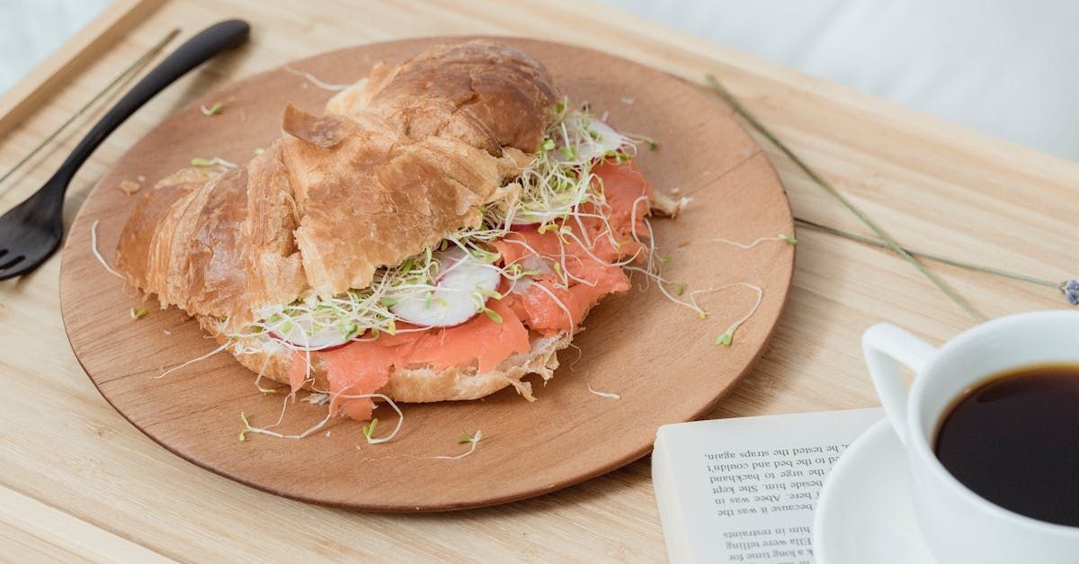 A sandwich on a croissant on a wooden plate next to a cup of coffee.