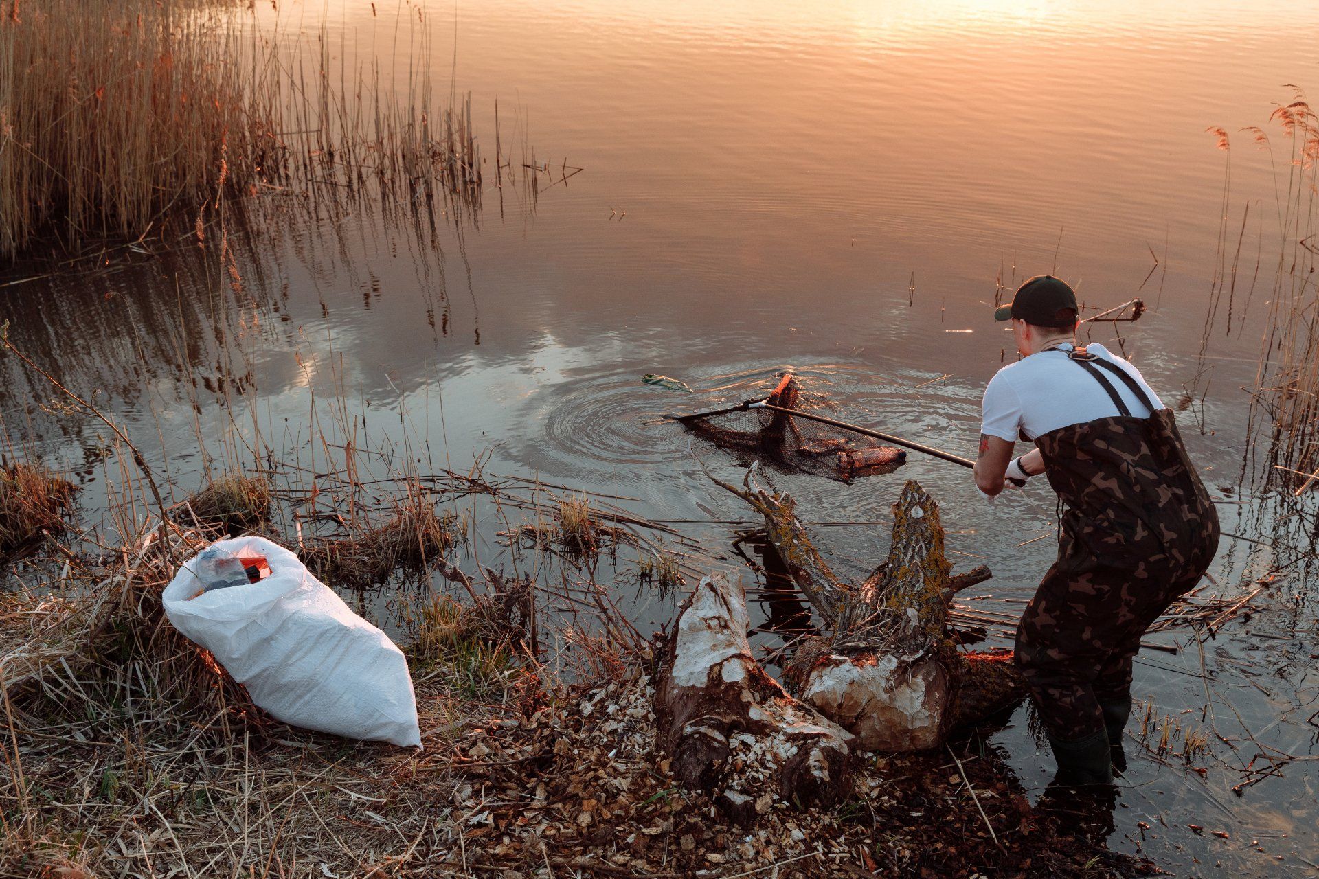 A man with hip waders on trying to fish trash out of a lake with a fishing pole