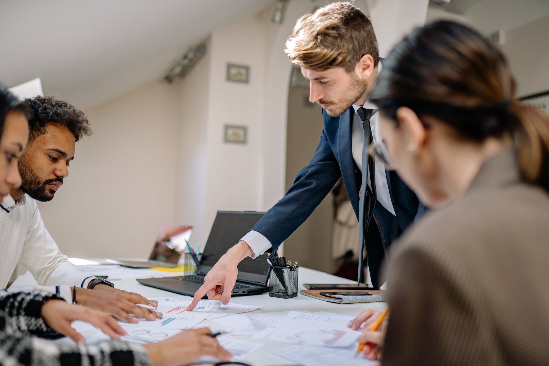 Business team in a meeting; man in suit points at document, others look on.