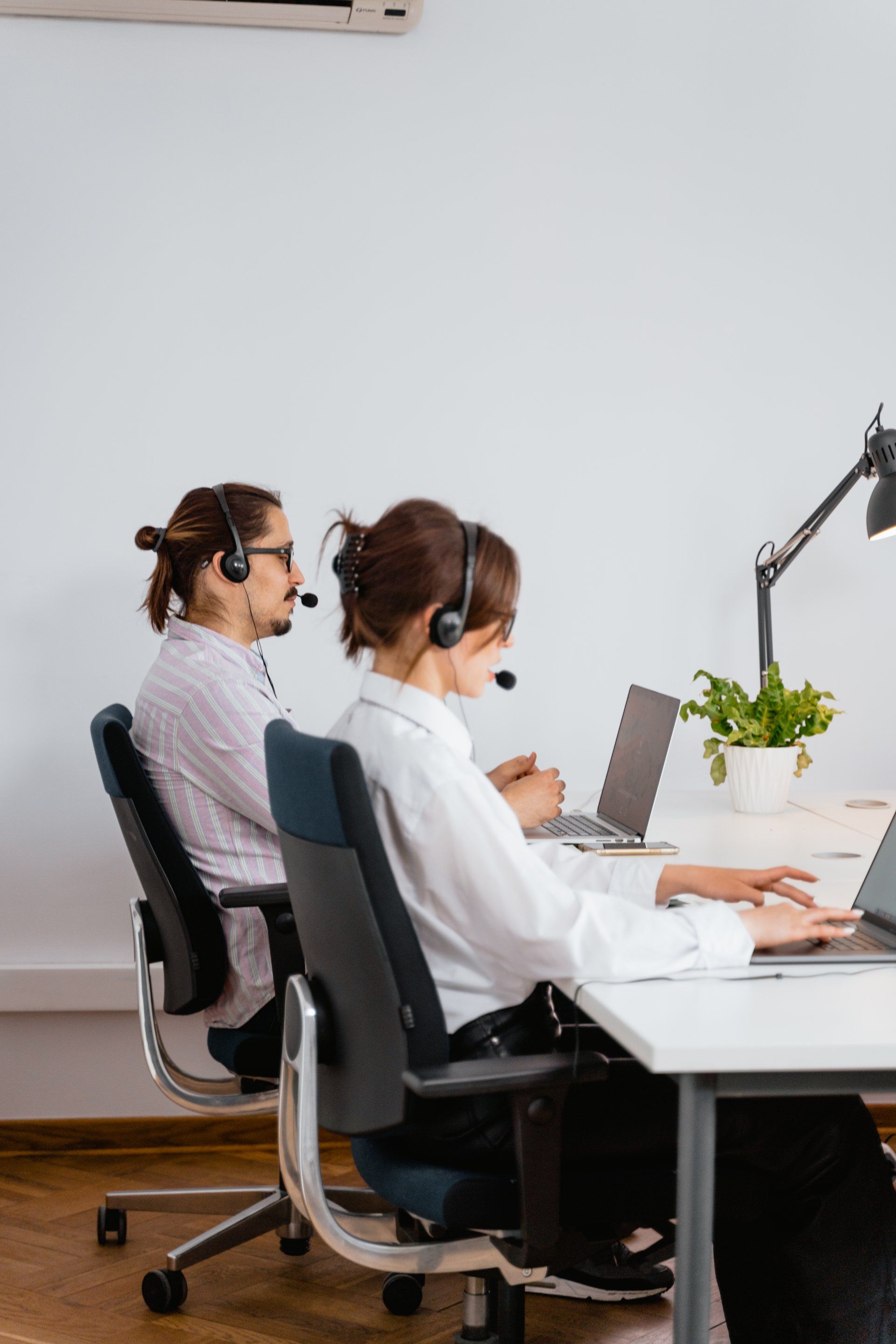 A man and a woman wearing headsets are sitting at a desk with laptops.