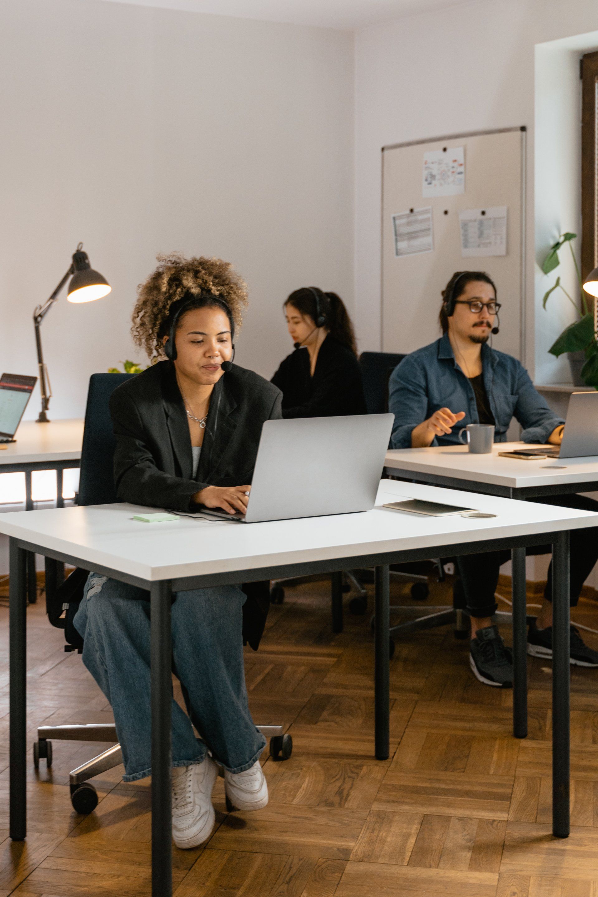 Three office workers wearing headsets use laptops at their desks. The woman in front has curly hair and is wearing jeans.