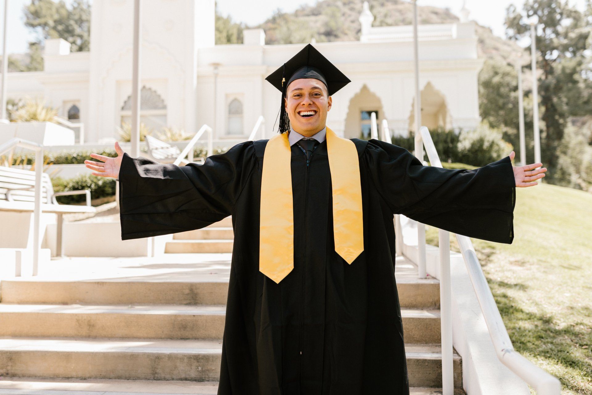A Young white man in black cap & gown with yellow valedictorian sash over his shoulders