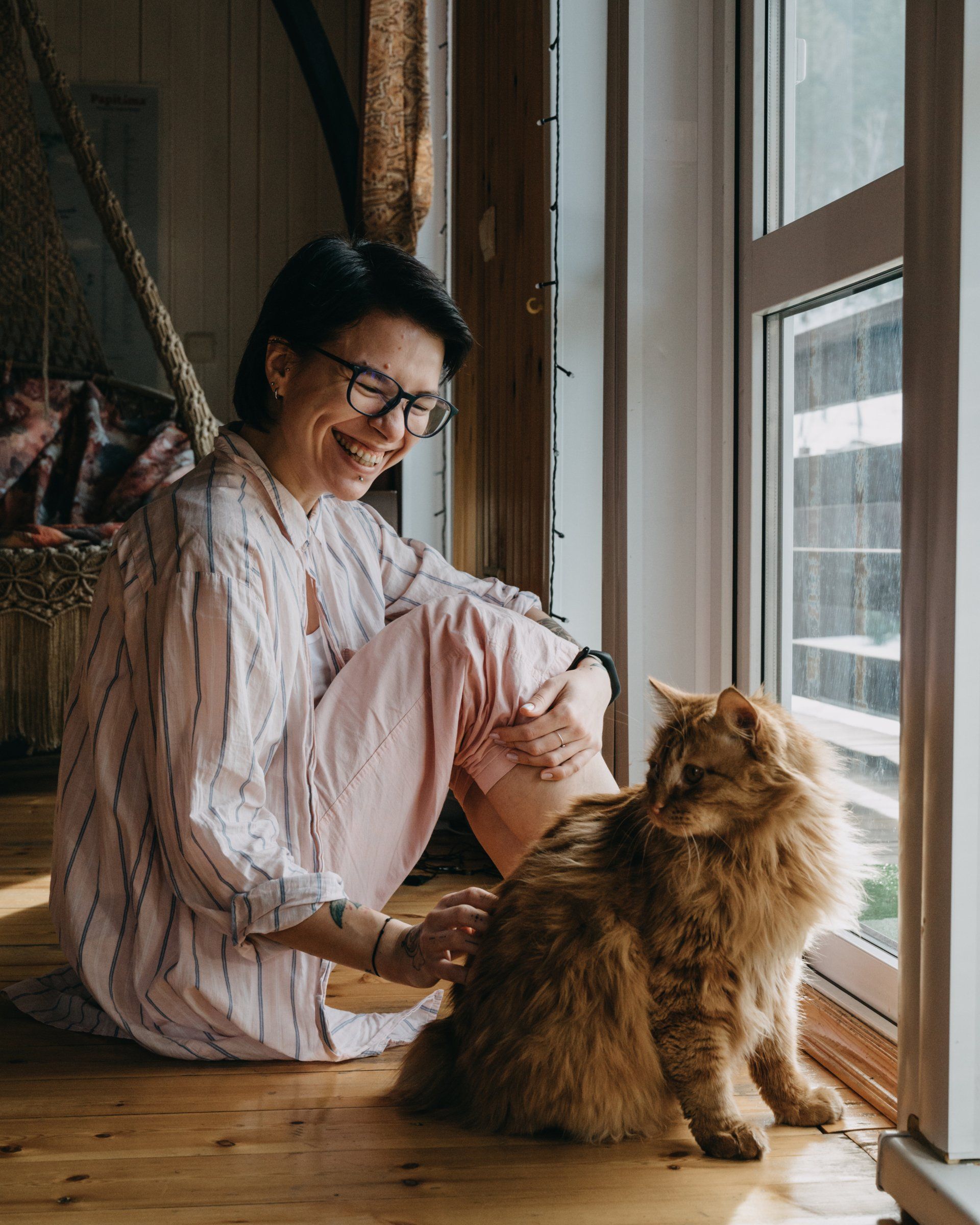 A woman is sitting on the floor with a cat in front of a window.