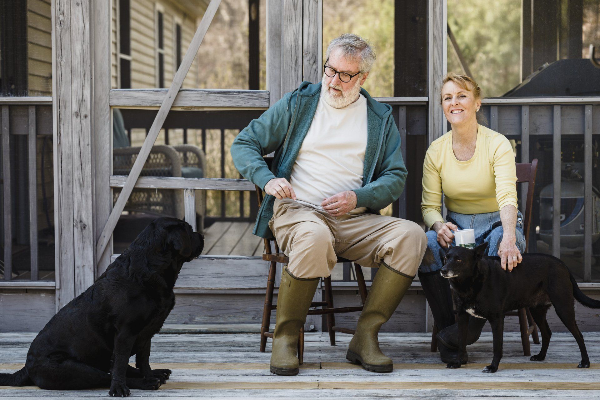 a man and a woman are sitting on a porch with their dogs .