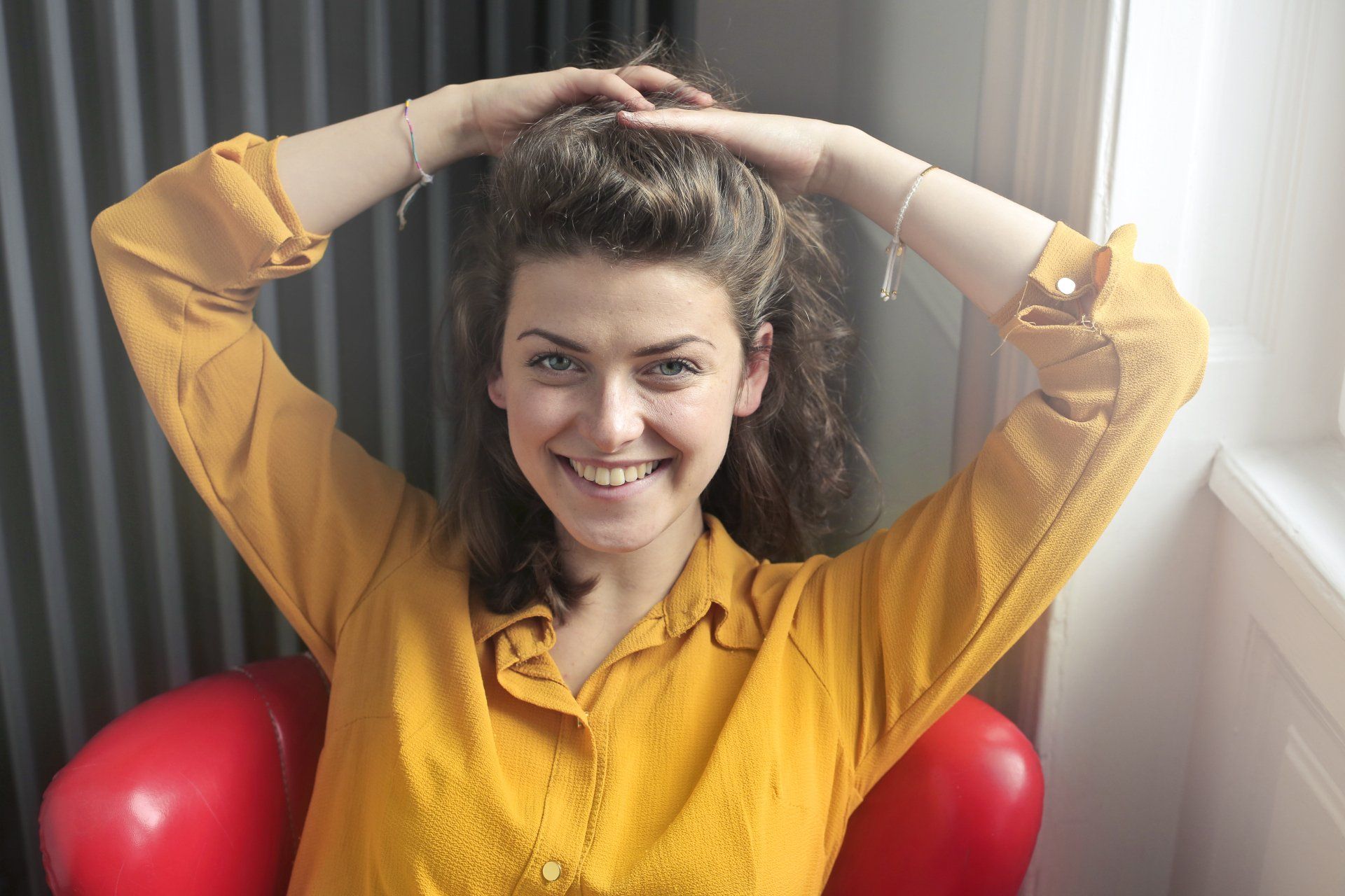 A woman in a yellow shirt is sitting in a red chair with her hands in her hair smiling.