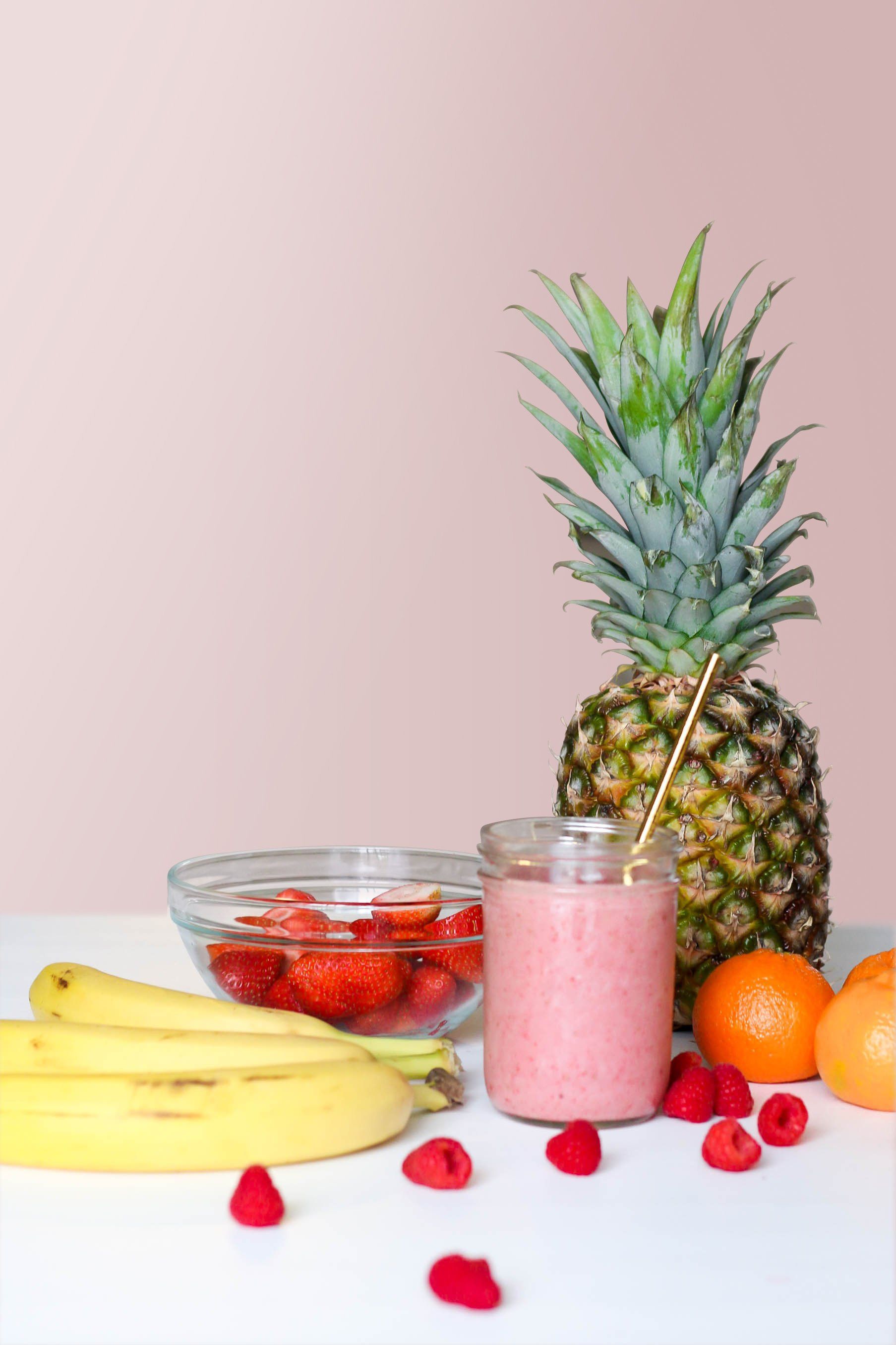 A smoothie is sitting on a table next to a pineapple , bananas , raspberries and oranges.
