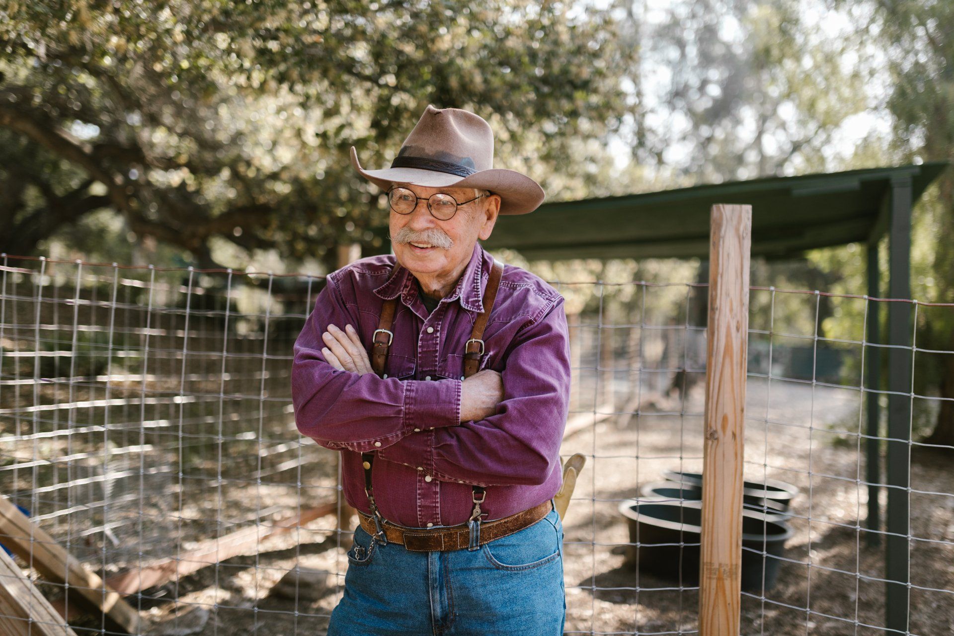 an older man wearing a cowboy hat and suspenders is standing in front of a fence .