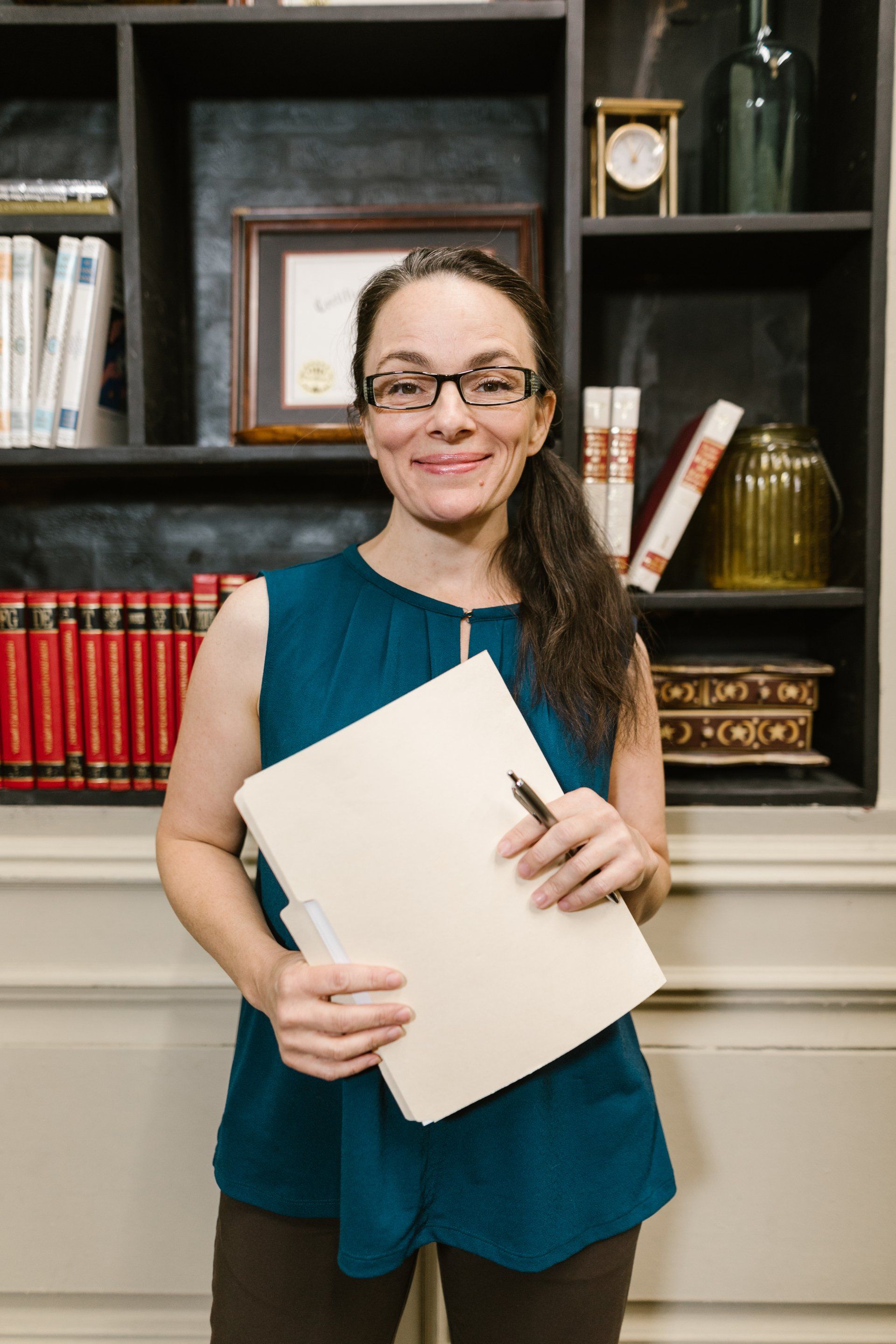A woman is holding a notebook and a pen in front of a bookshelf.