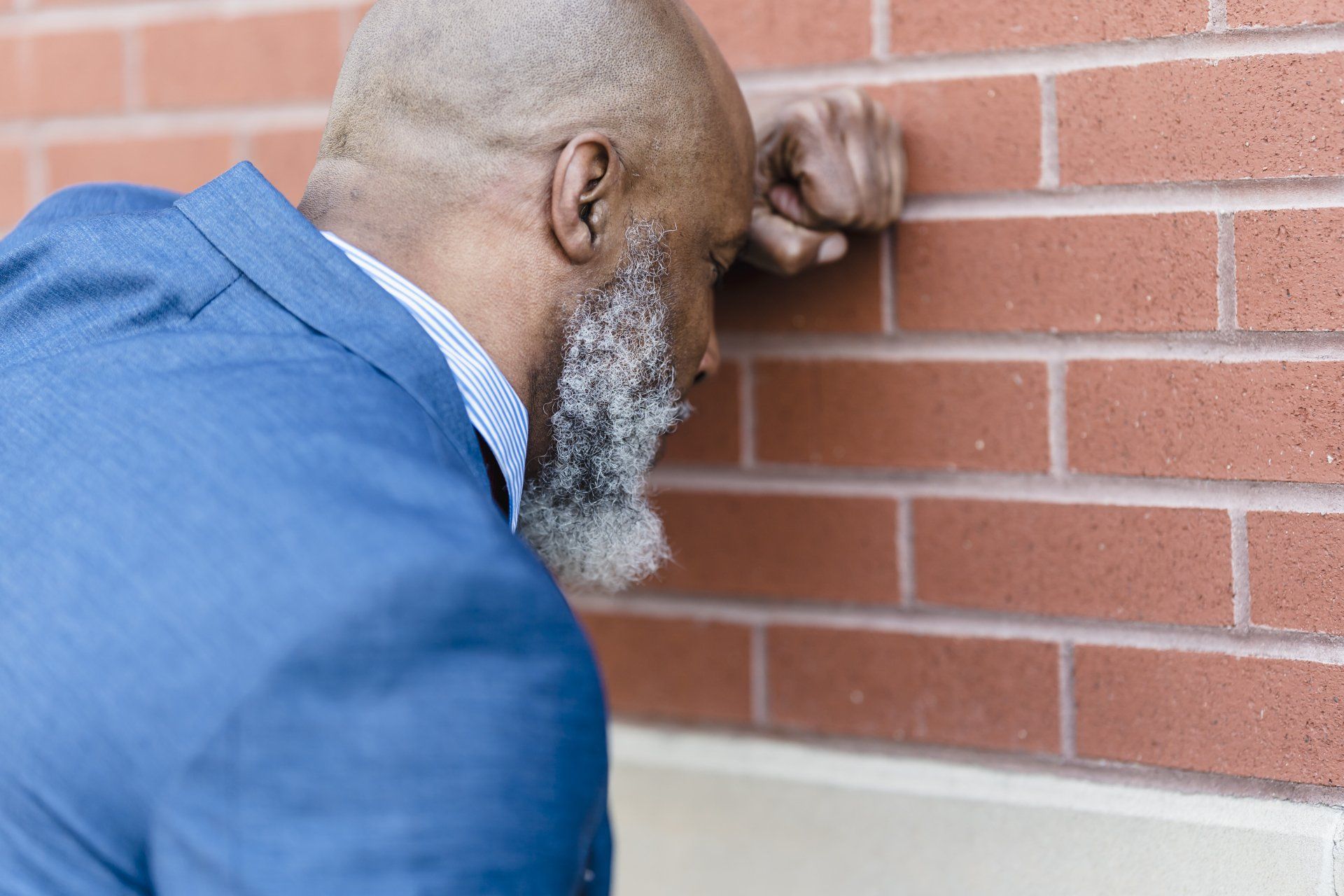 Black man with salt and pepper beard and blue suit, head on fist up against brick wall